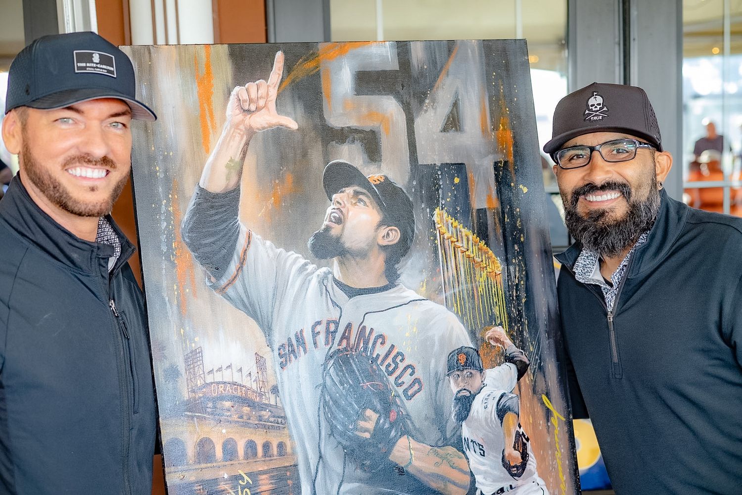 Two men smiling beside a painted portrait of a San Francisco Giants player wearing number 54.