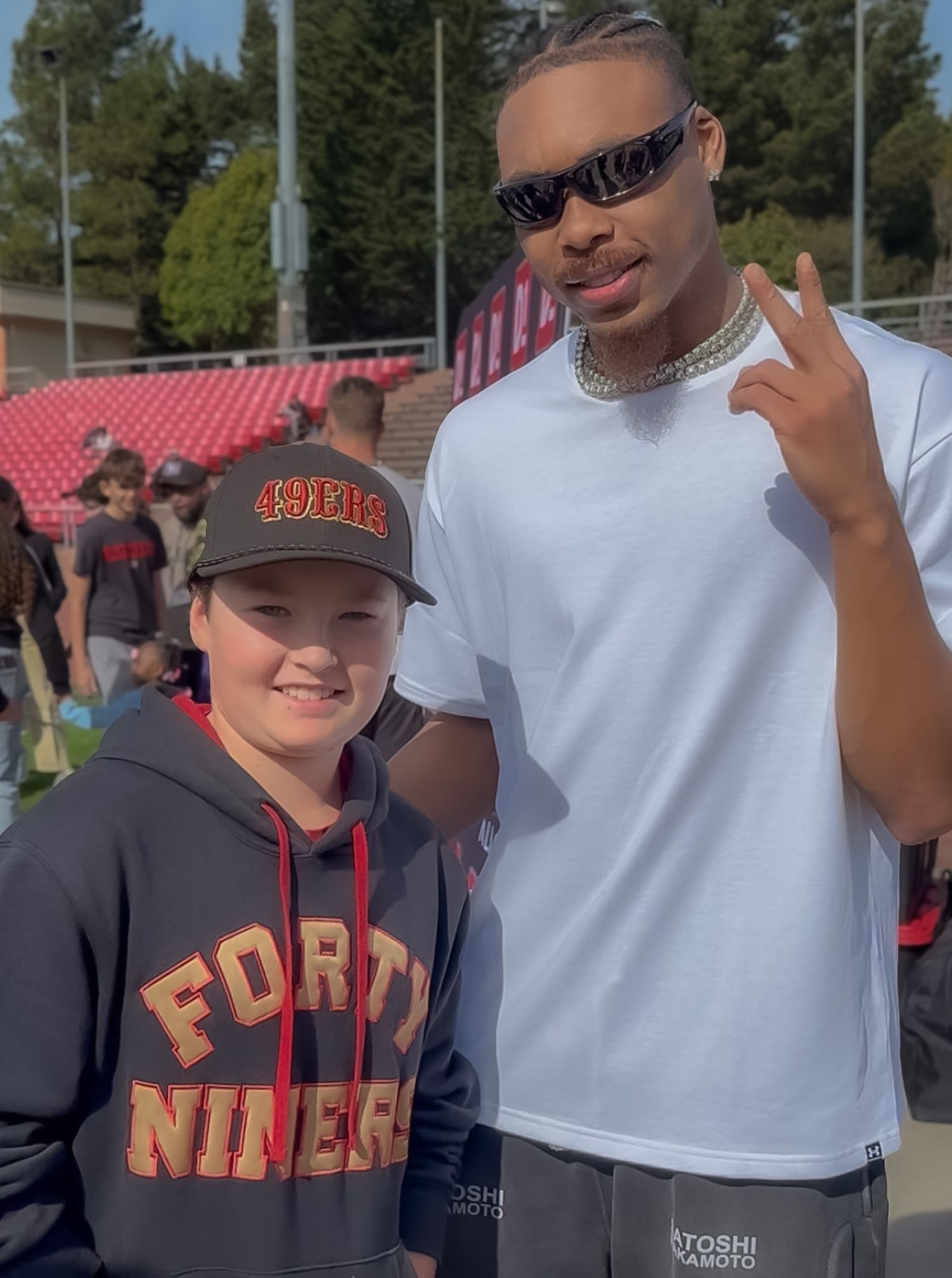 Young fan in 49ers hoodie and cap poses with Justin Jefferson in white tee and sunglasses at outdoor event.