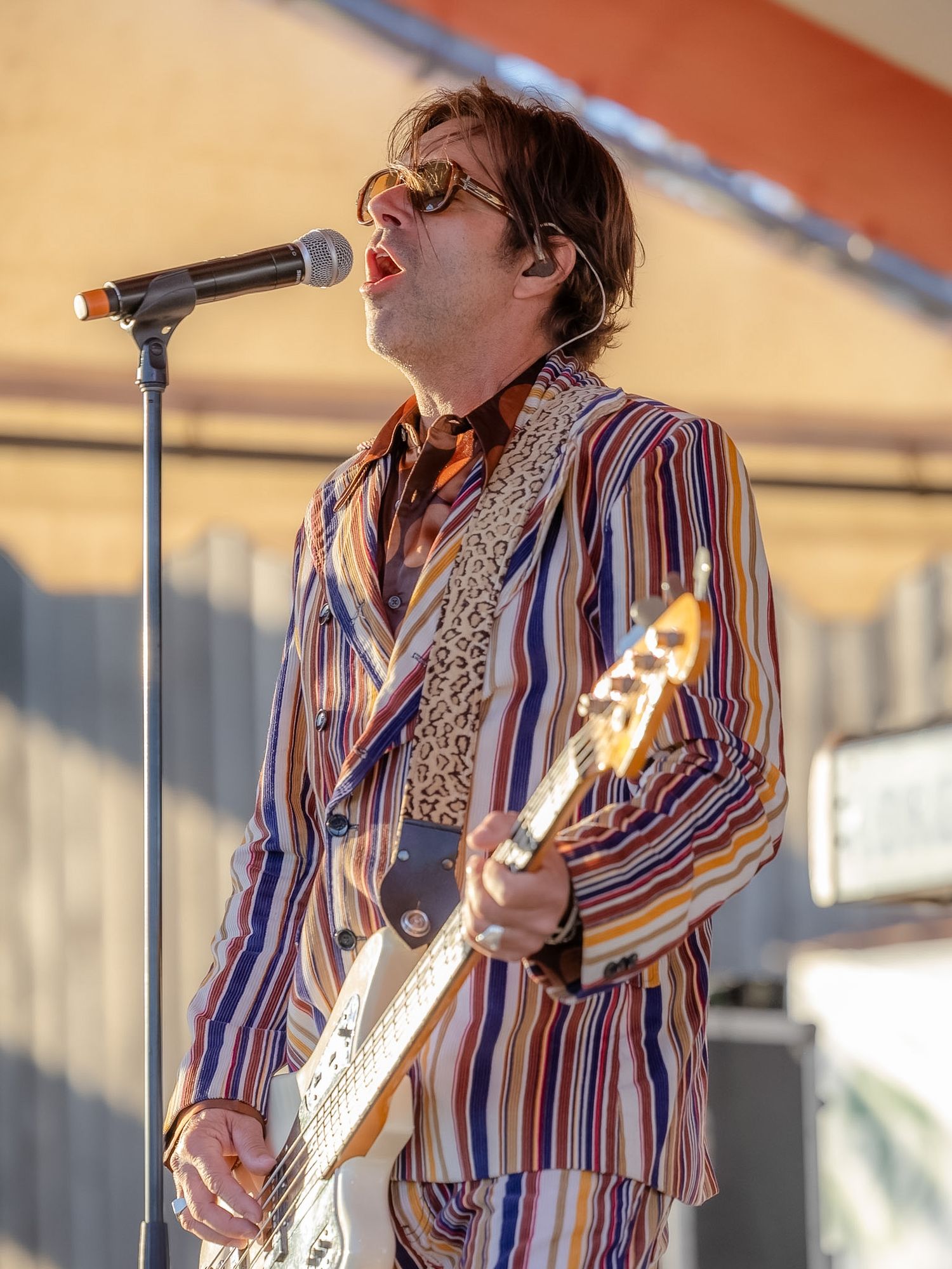 Male musician in striped suit playing electric guitar and singing into microphone at outdoor concert.
