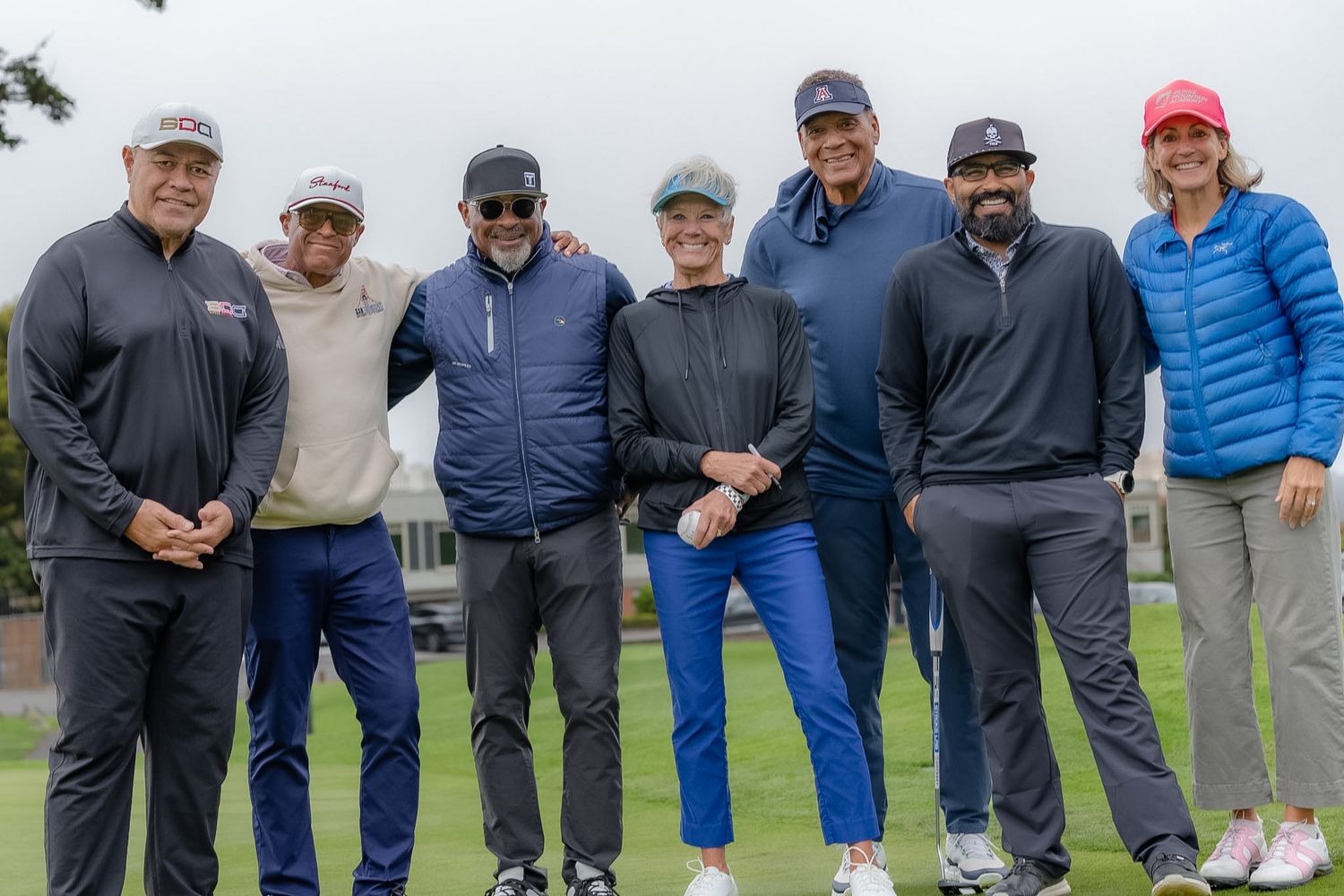 Seven people pose together smiling on a golf course on an overcast day, dressed in casual athletic wear.