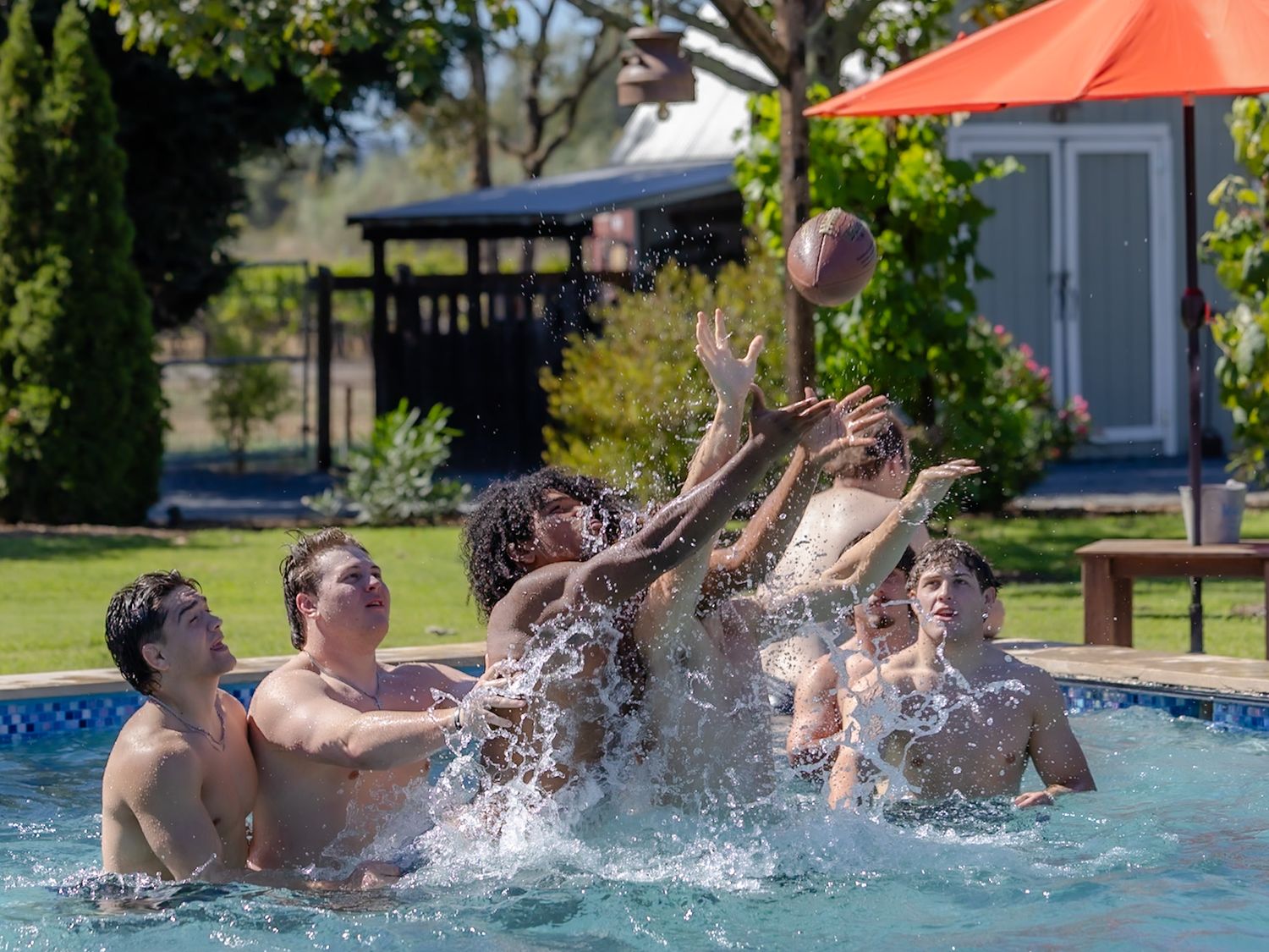 Group of teens splashing in a backyard pool playing with a football on a sunny day.