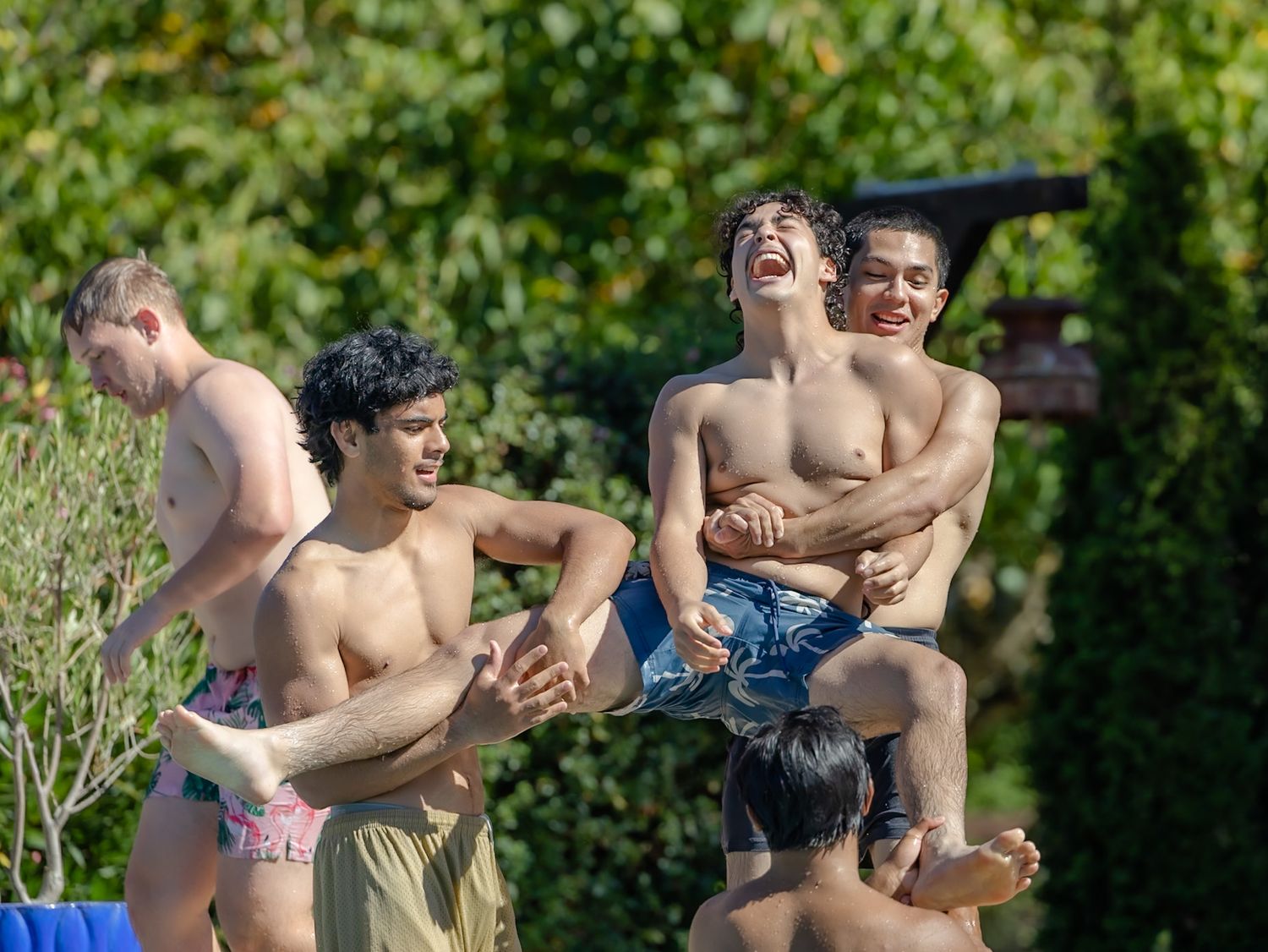 Group of shirtless young men laughing and playing by a pool, lifting each other up outdoors.