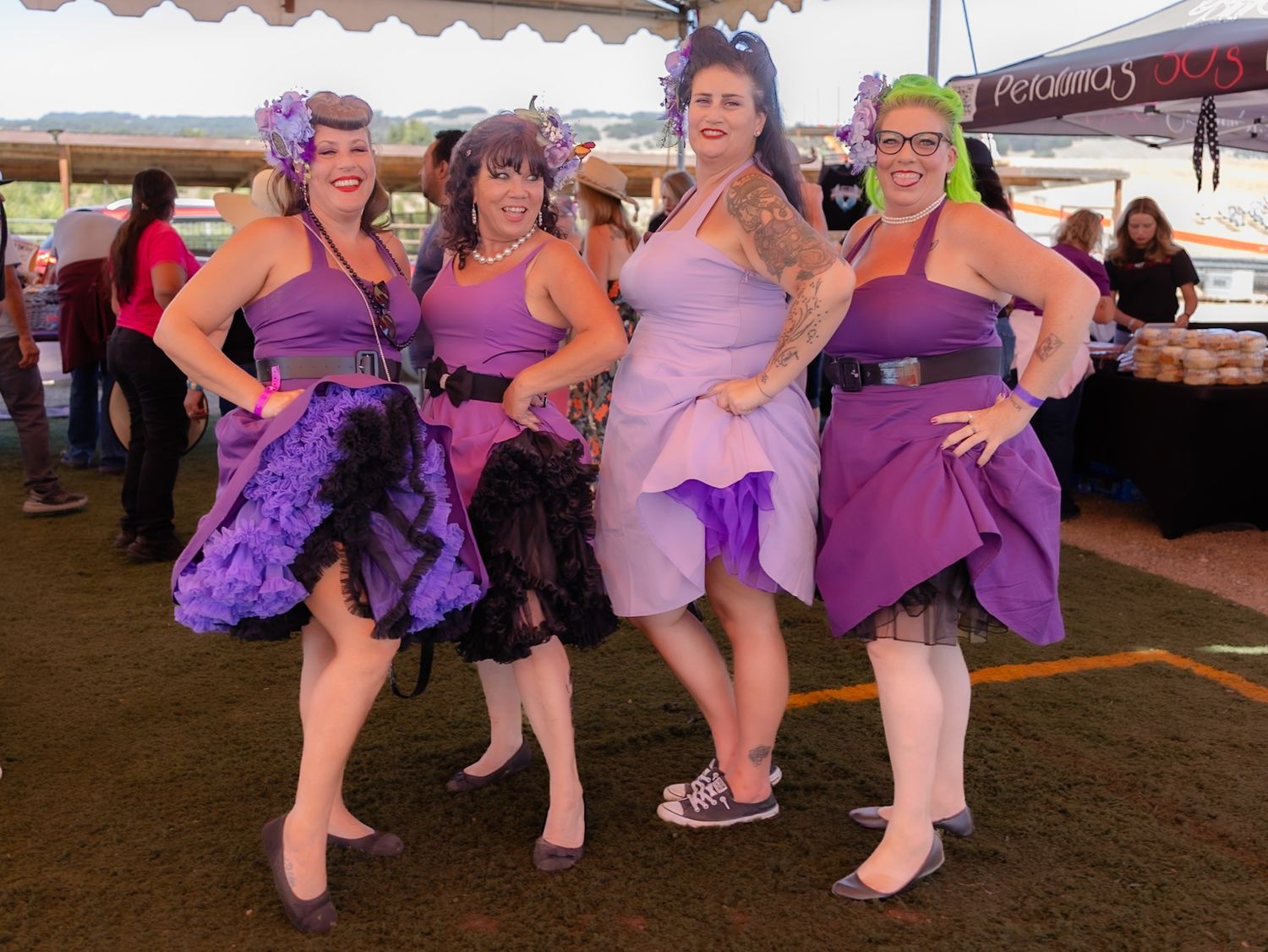 Four women in purple dresses pose together at an outdoor event under a tent.