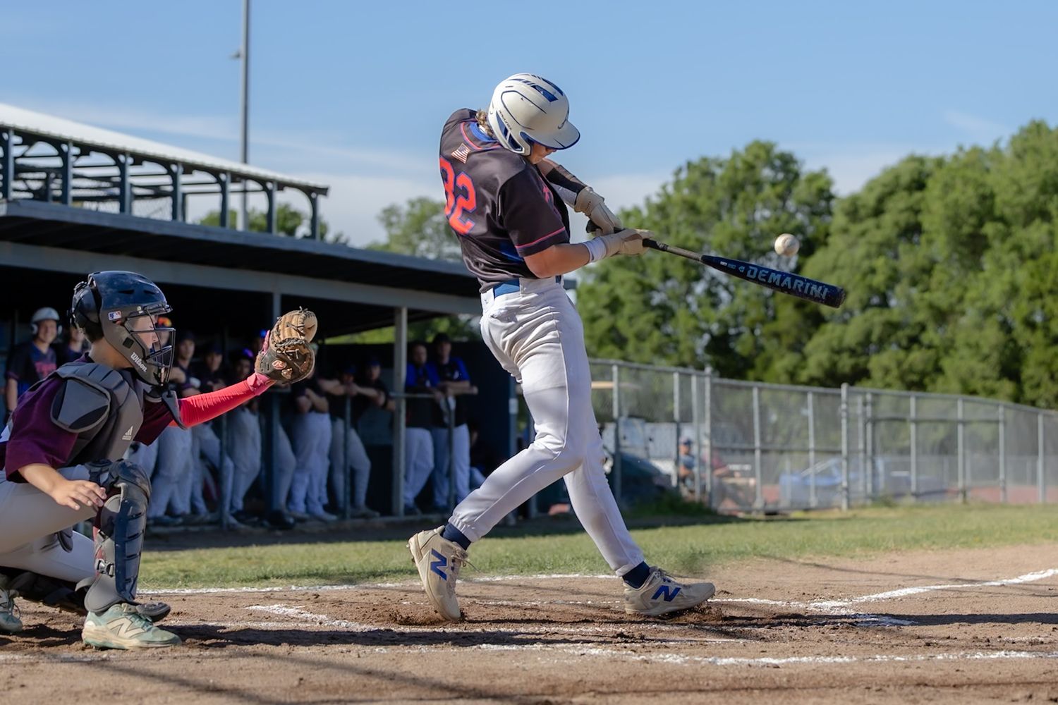 Baseball batter swinging at pitch during game, catcher crouching behind home plate, dugout with players visible in background.