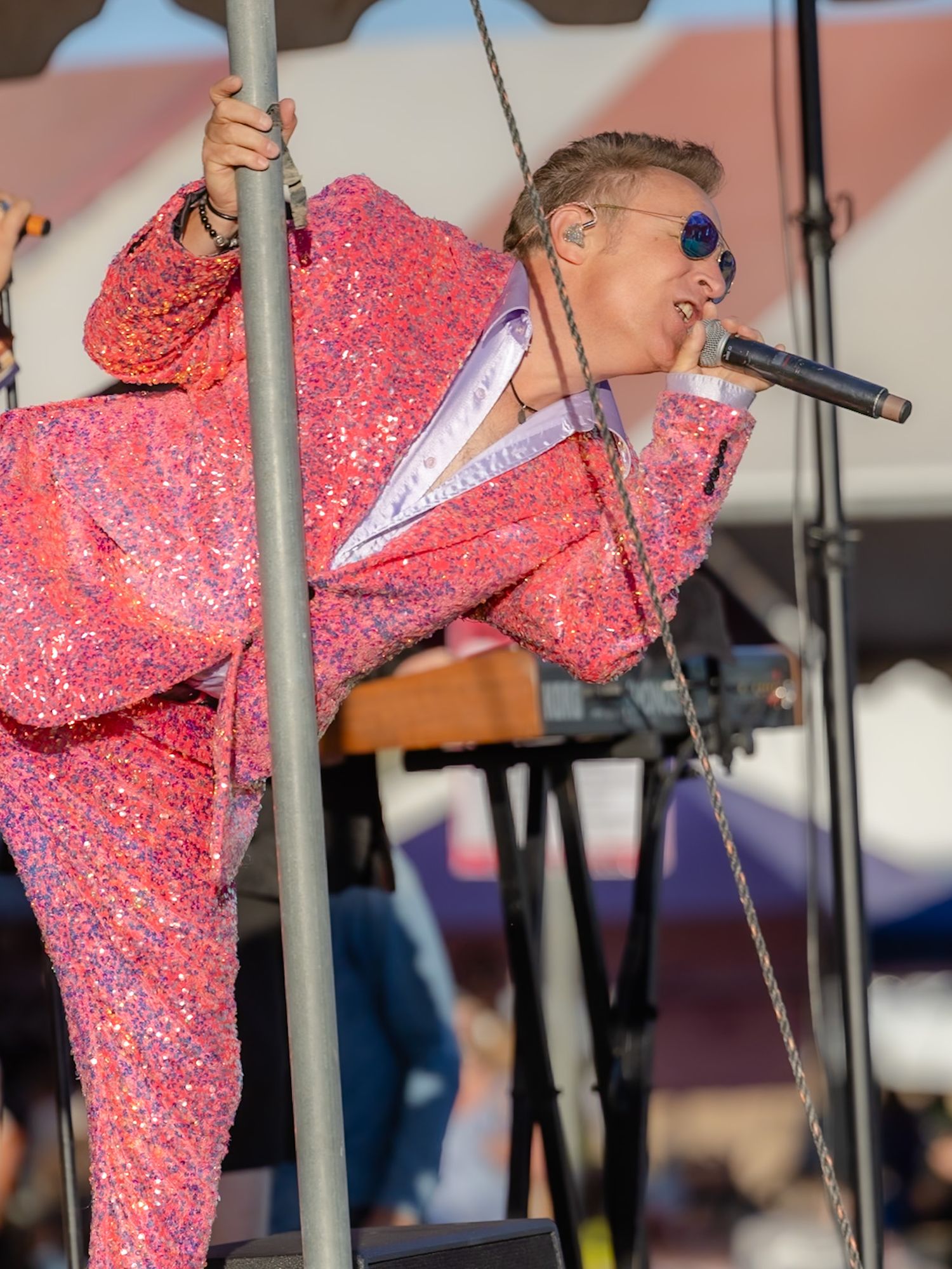Performer in pink sequin suit singing on stage, leaning forward with microphone at outdoor music event.