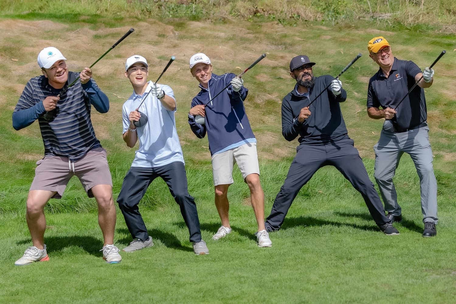 Five golfers pose playfully on a golf course, holding clubs in fun stances on a sunny day.