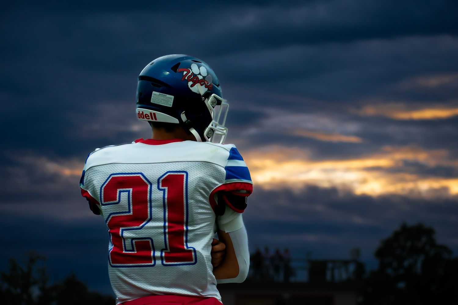 Football player wearing jersey number 21 stands on field at dusk under dramatic cloudy sky.
