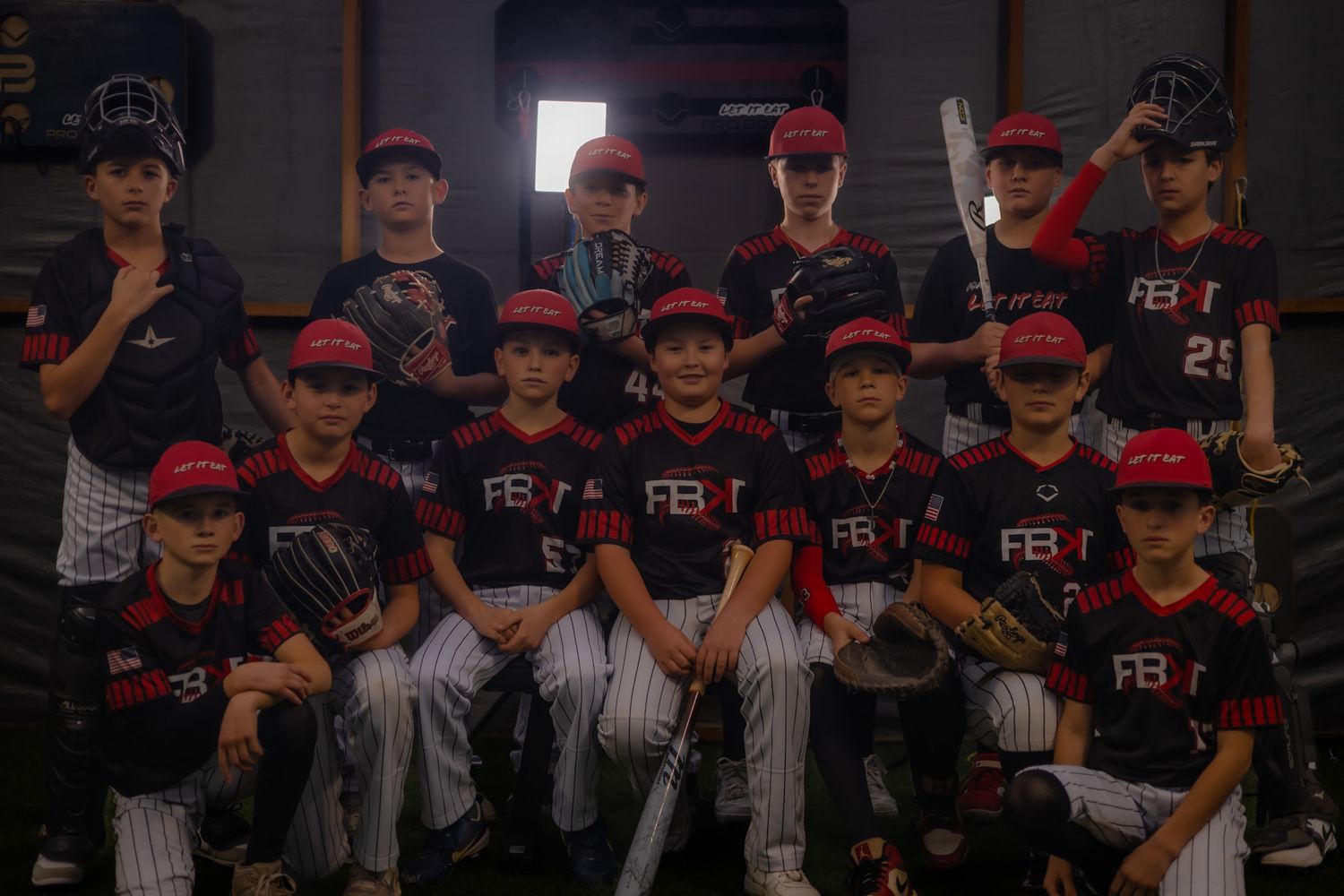 Youth baseball team in black and red uniforms posing together indoors holding bats and gloves for a team photo.