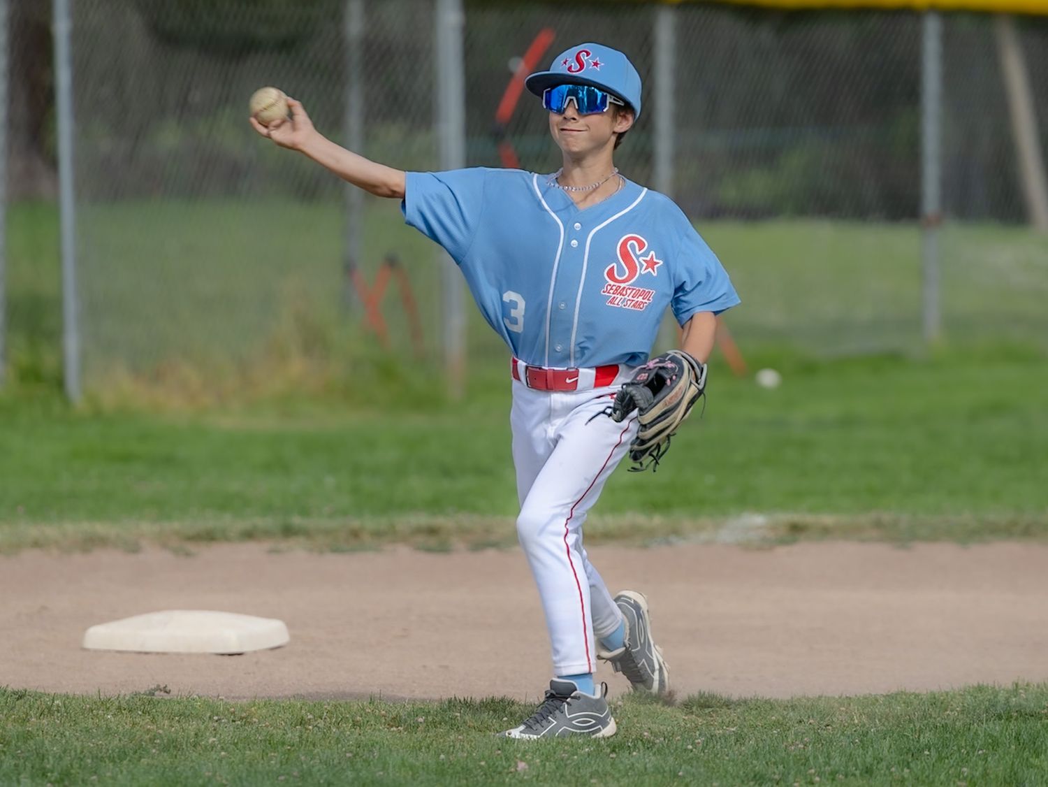 Youth baseball player in blue jersey throwing ball from second base during a sunny outdoor game.