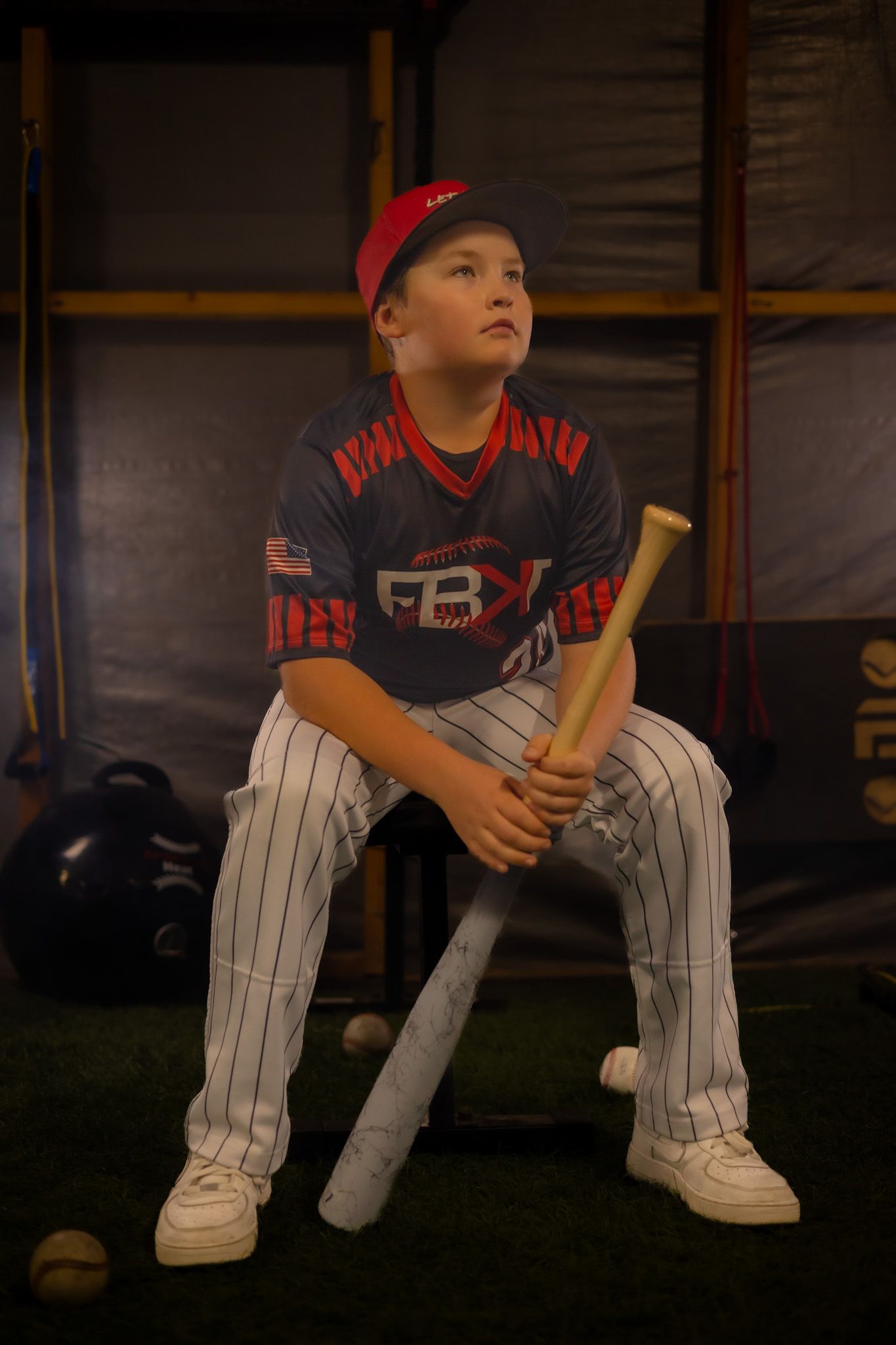 Young baseball player in FBX uniform sits holding a bat in a dugout, wearing a red cap and pinstripe pants.