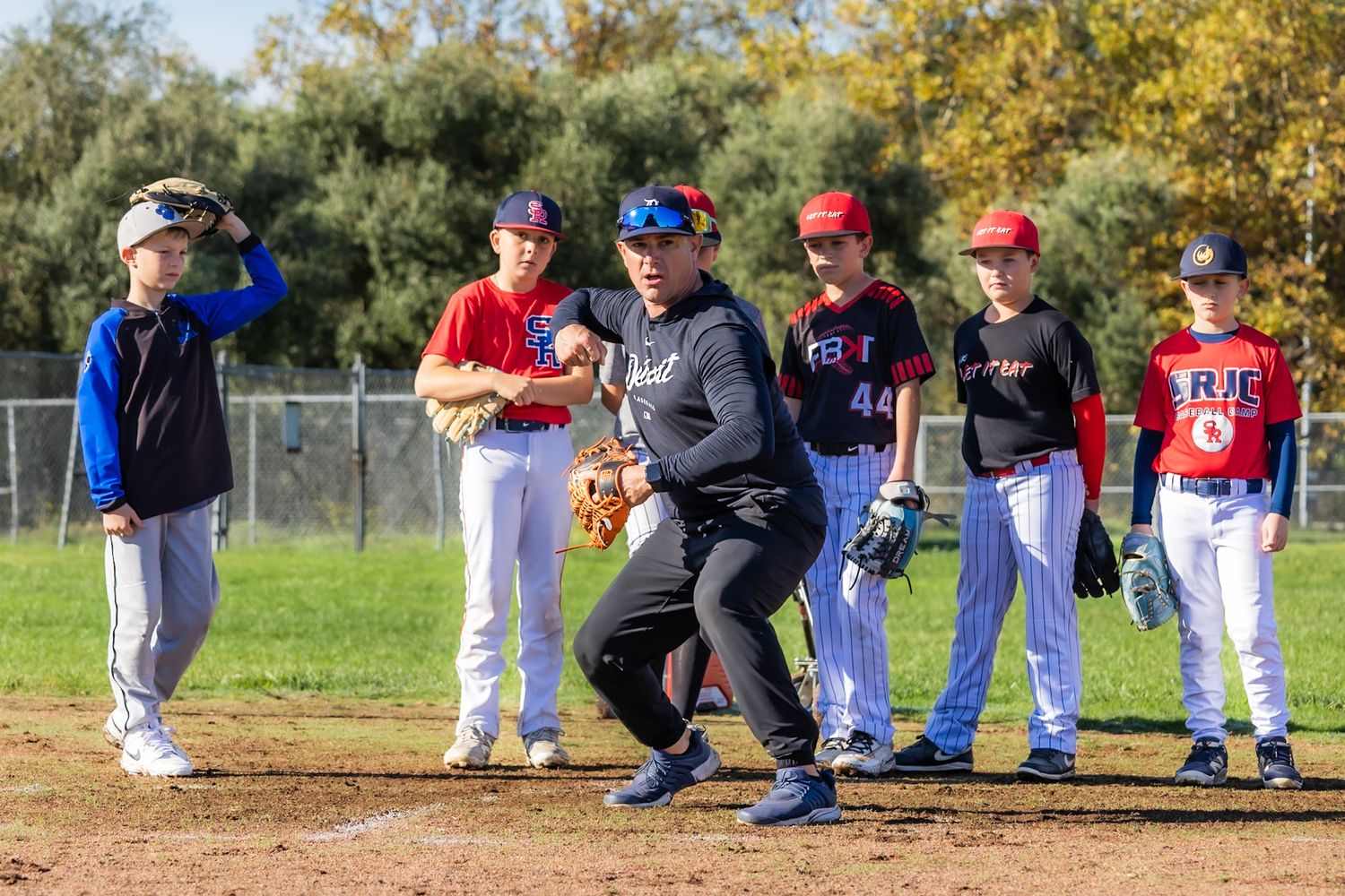 Adult baseball coach demonstrating fielding stance to youth players in red and blue uniforms during outdoor practice.