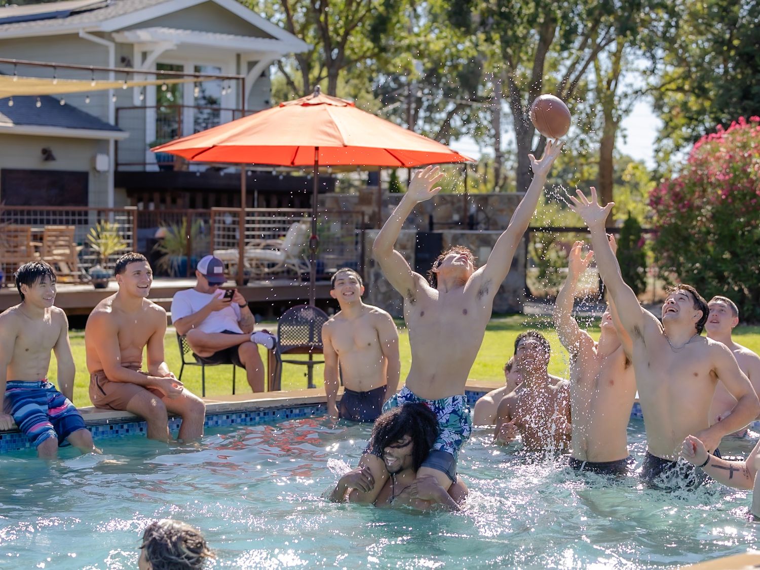 Group of teens playing football in a backyard pool on a sunny day with an orange patio umbrella in the background.