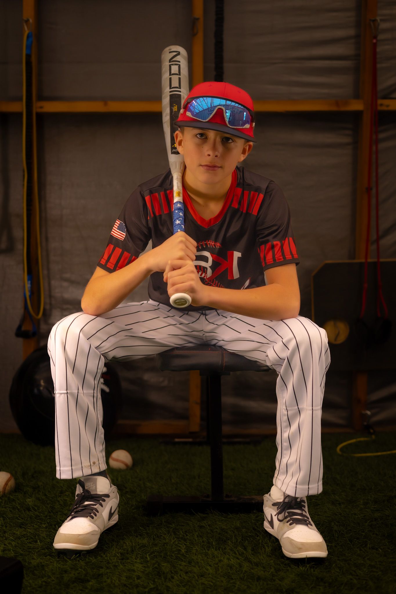 Young baseball player in uniform holds bat while seated, wearing red cap and blue sunglasses in dugout setting.