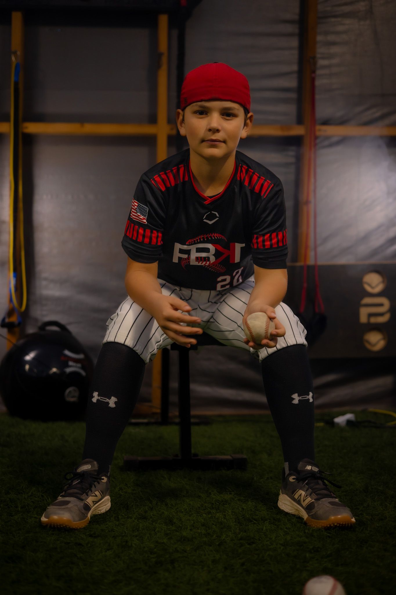Young baseball player in FBH jersey and red cap sits holding a baseball in an indoor training facility.