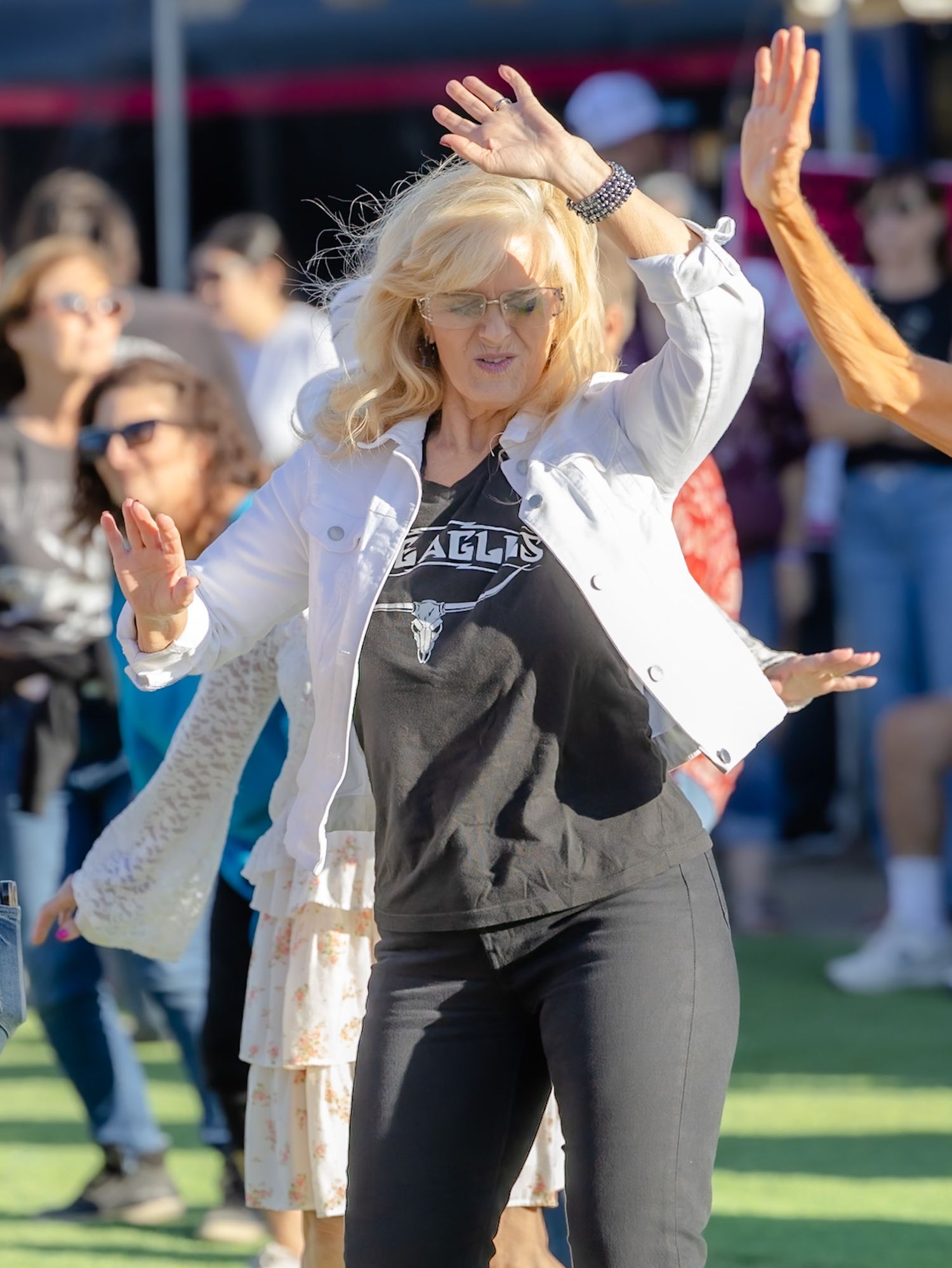 Blonde woman in Eagles band tee and white jacket dancing outdoors at a sunny event with hands raised.
