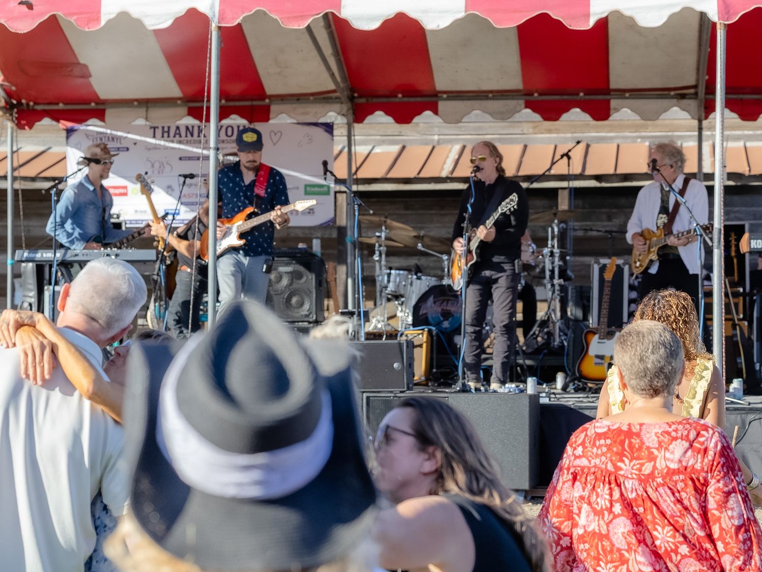 Live band performing on outdoor stage under red and white tent canopy for crowd of spectators at daytime festival.