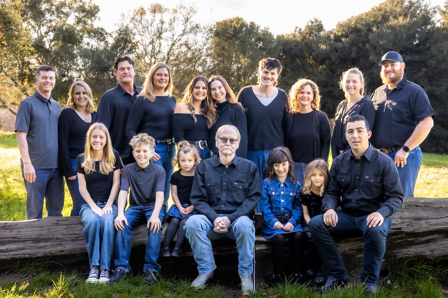 Large family group portrait outdoors, posing together near a rustic log in a park setting with green trees in background.