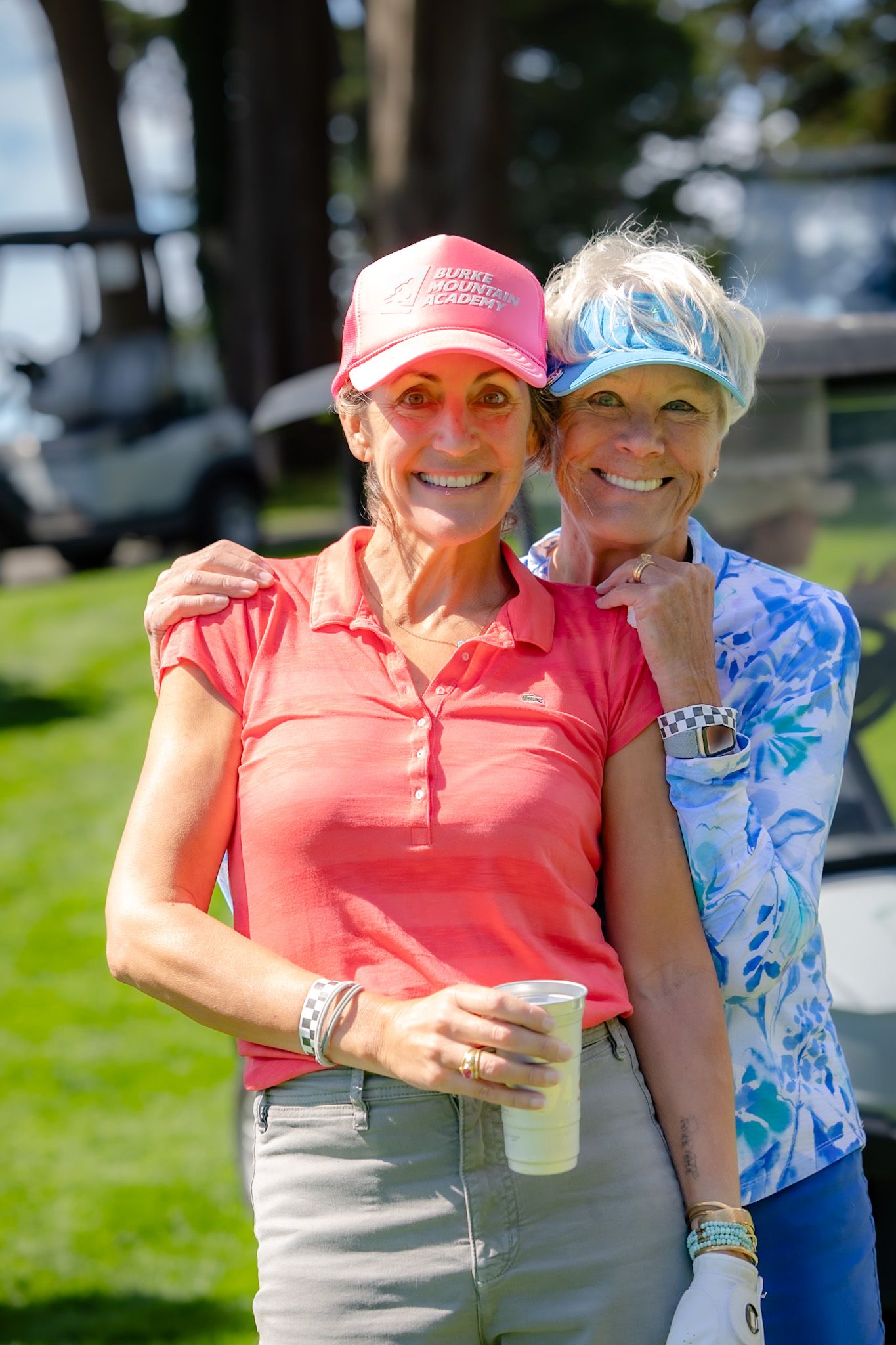 Two smiling women in golf attire and caps pose together outdoors on a sunny golf course.