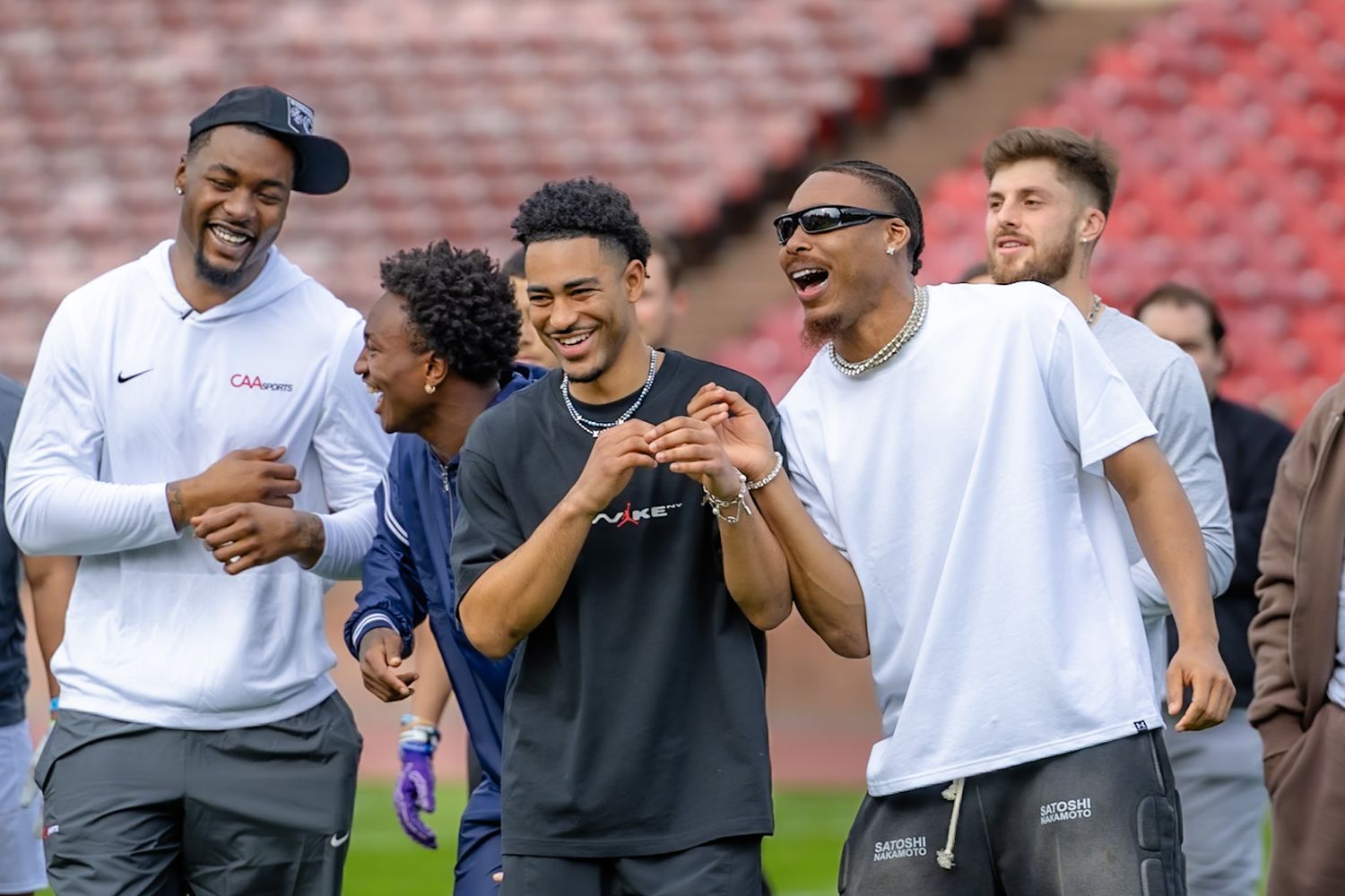 Group of young men laughing and socializing on a football field stadium, wearing casual streetwear clothing. Justin Jefferson Zae Flowers