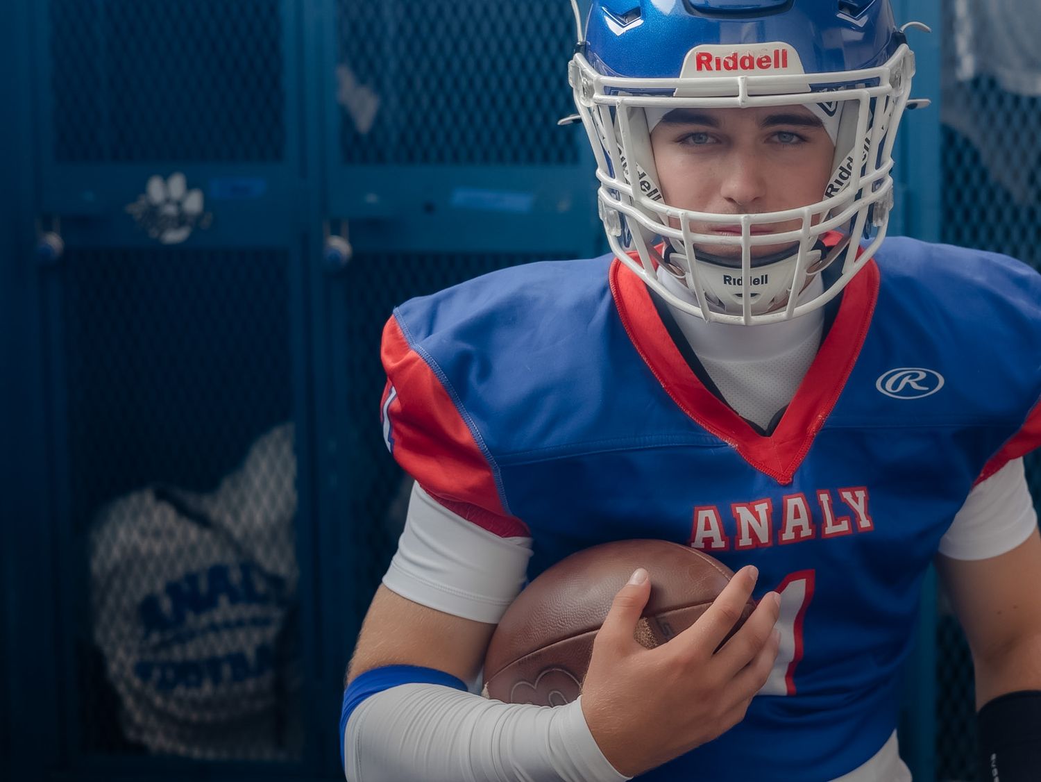 Analy high school football player in blue Riddell helmet holding a ball near blue lockers.
