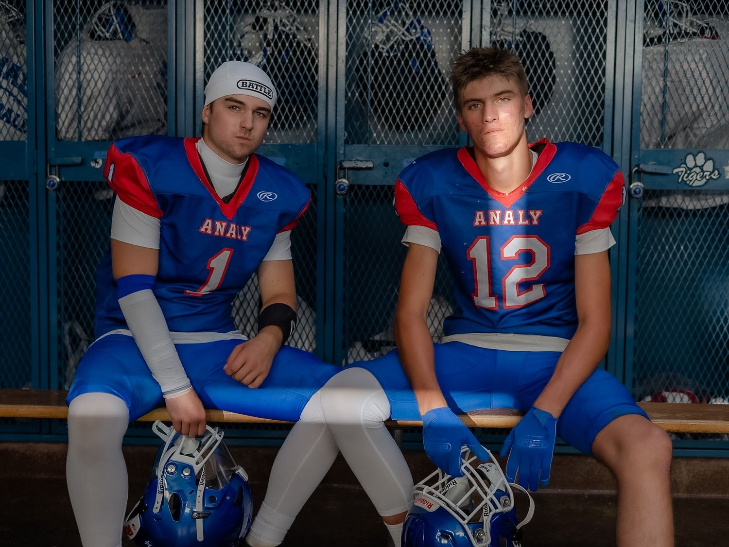 Two Analy High School football players in blue and red uniforms sit in locker room benches holding helmets.