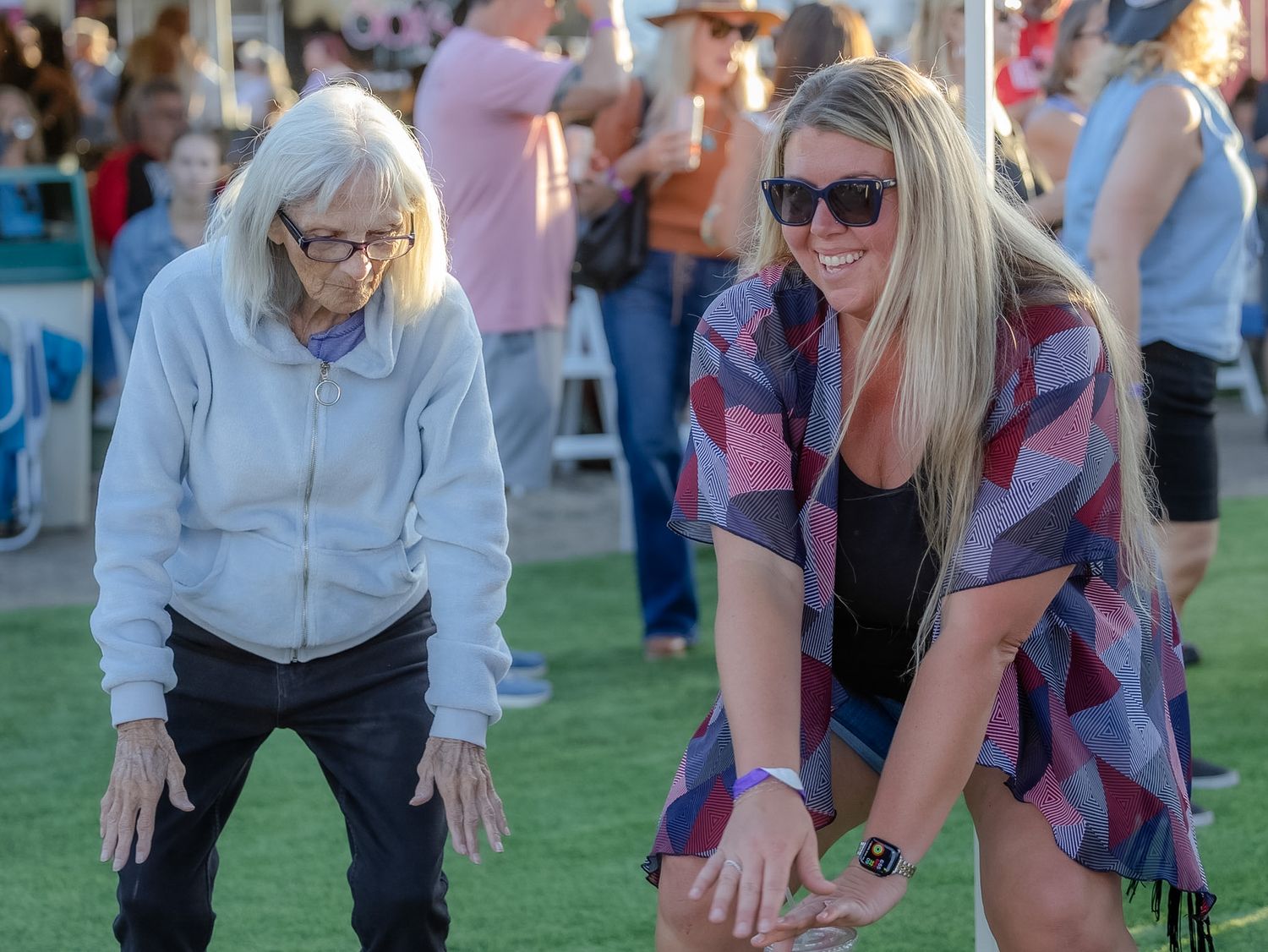 Two women playing bocce ball on a green lawn at an outdoor event, one elderly in gray, one blonde in plaid.