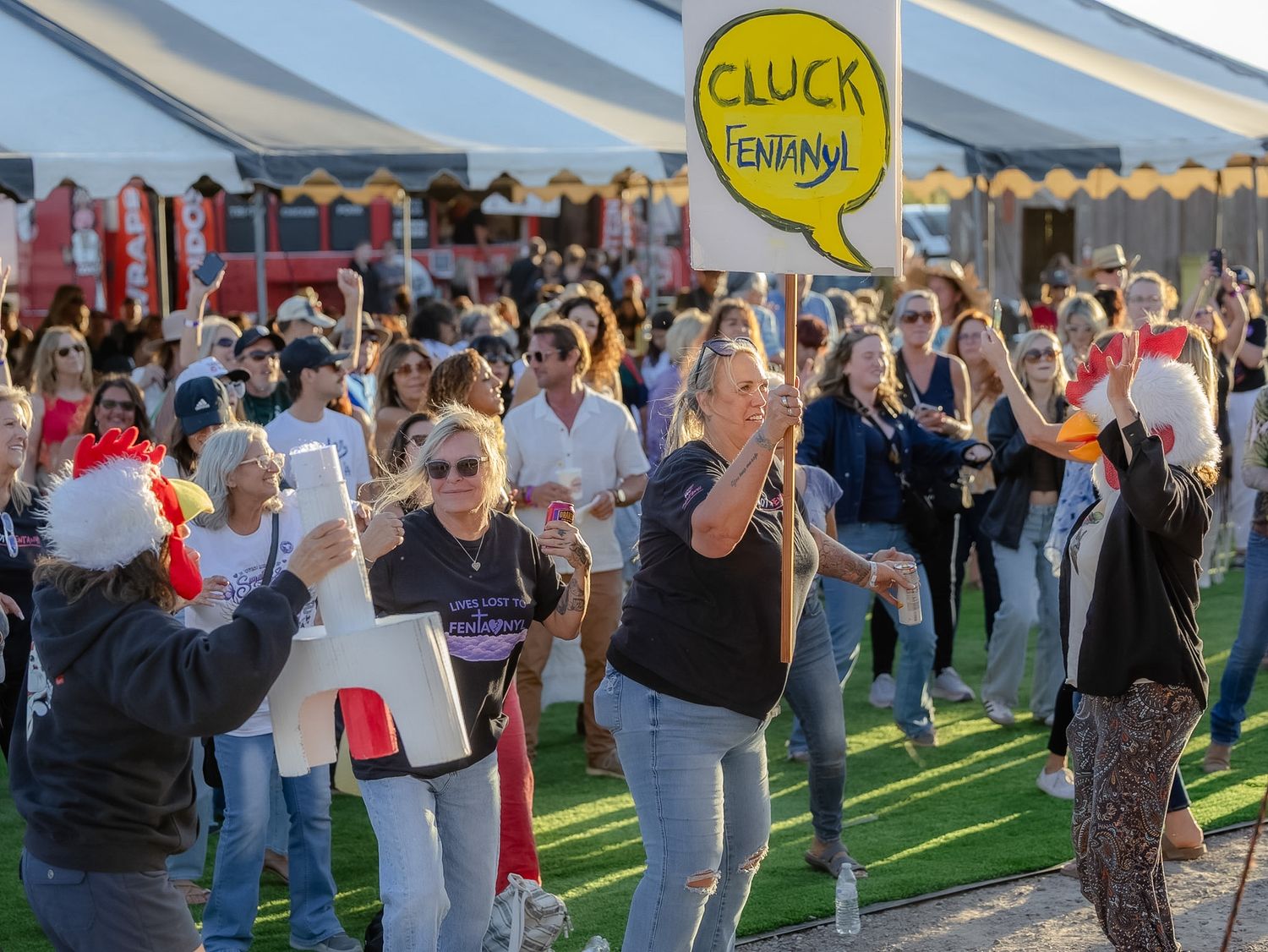 Crowd of women dancing outdoors at a festival event, with a yellow sign reading Cluck Henfany visible.
