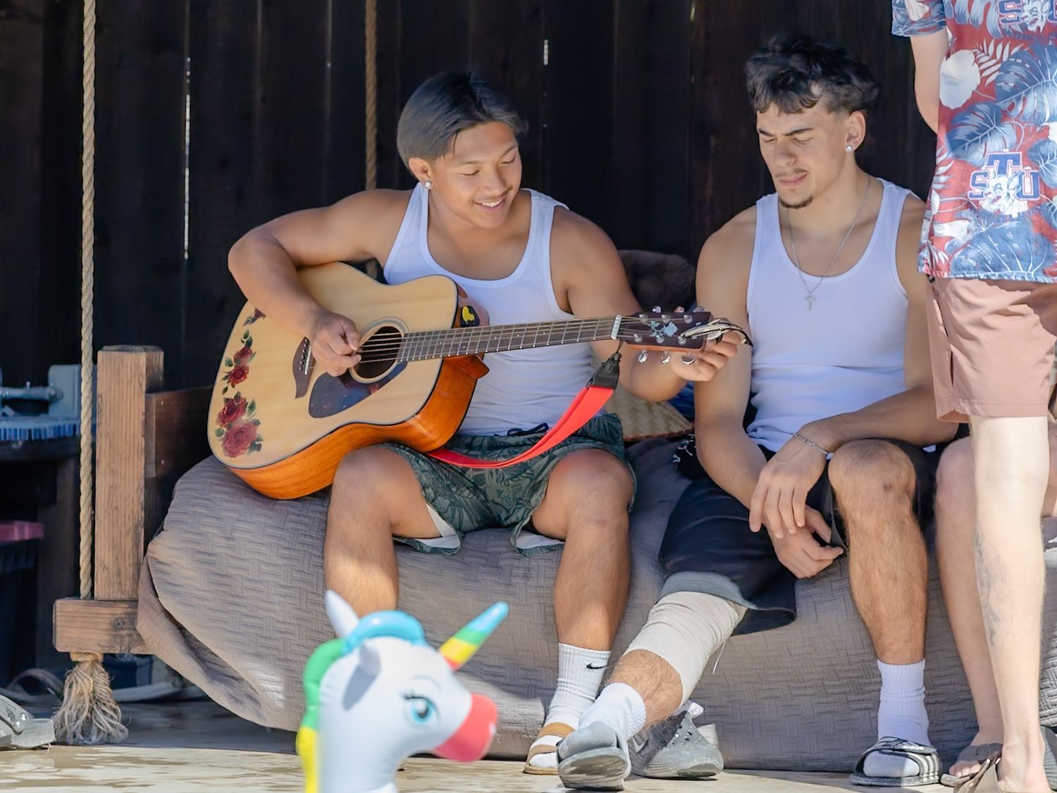 Two young men sitting outdoors, one playing an acoustic guitar, with a colorful unicorn toy on the ground.