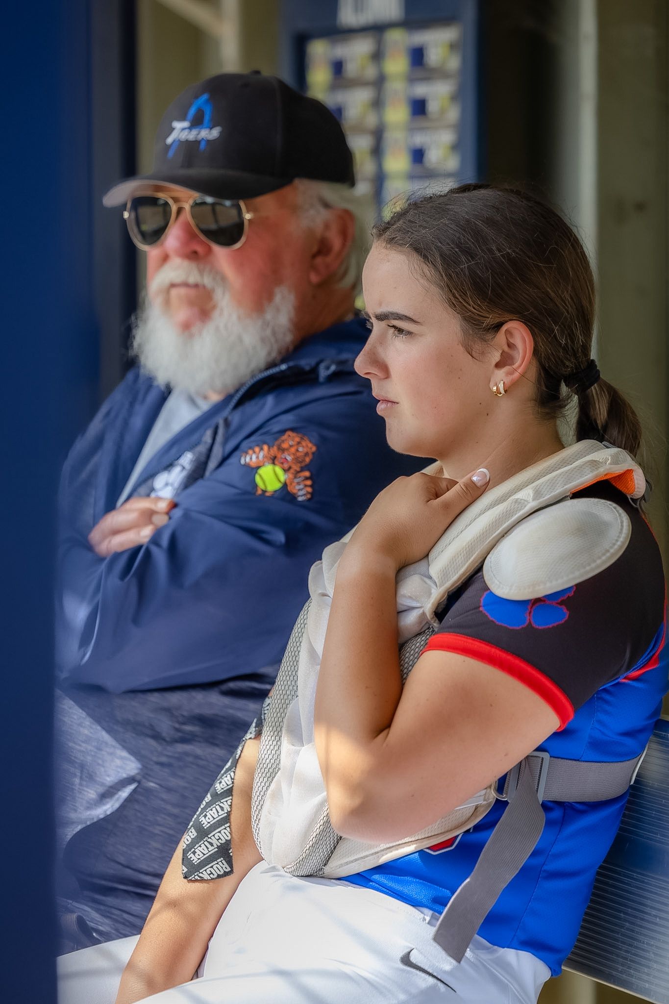 Young female softball catcher in dugout with older bearded coach wearing blue Twins cap during game.