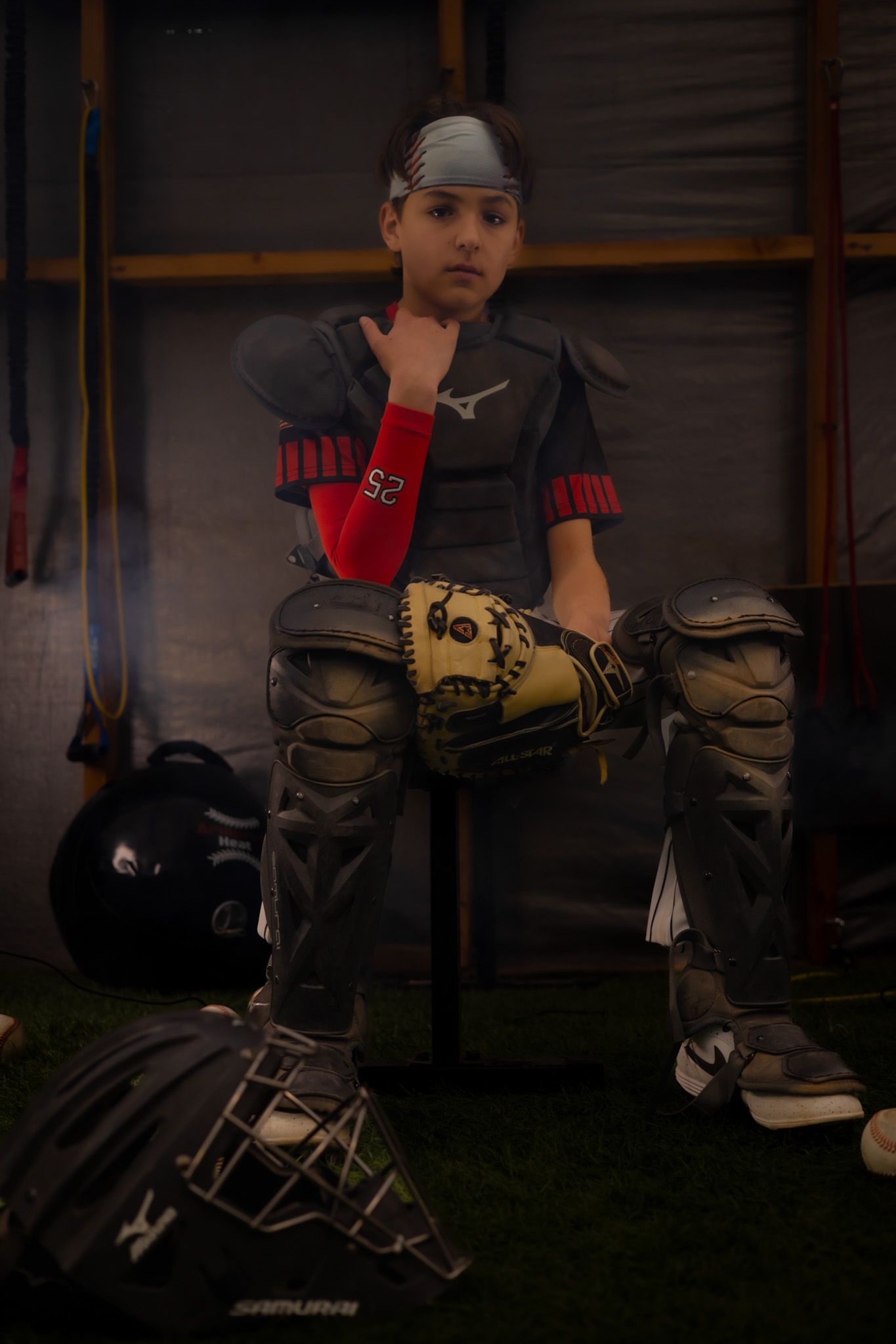 Young baseball catcher in full gear including chest protector, knee guards, and mitt sitting in dugout.