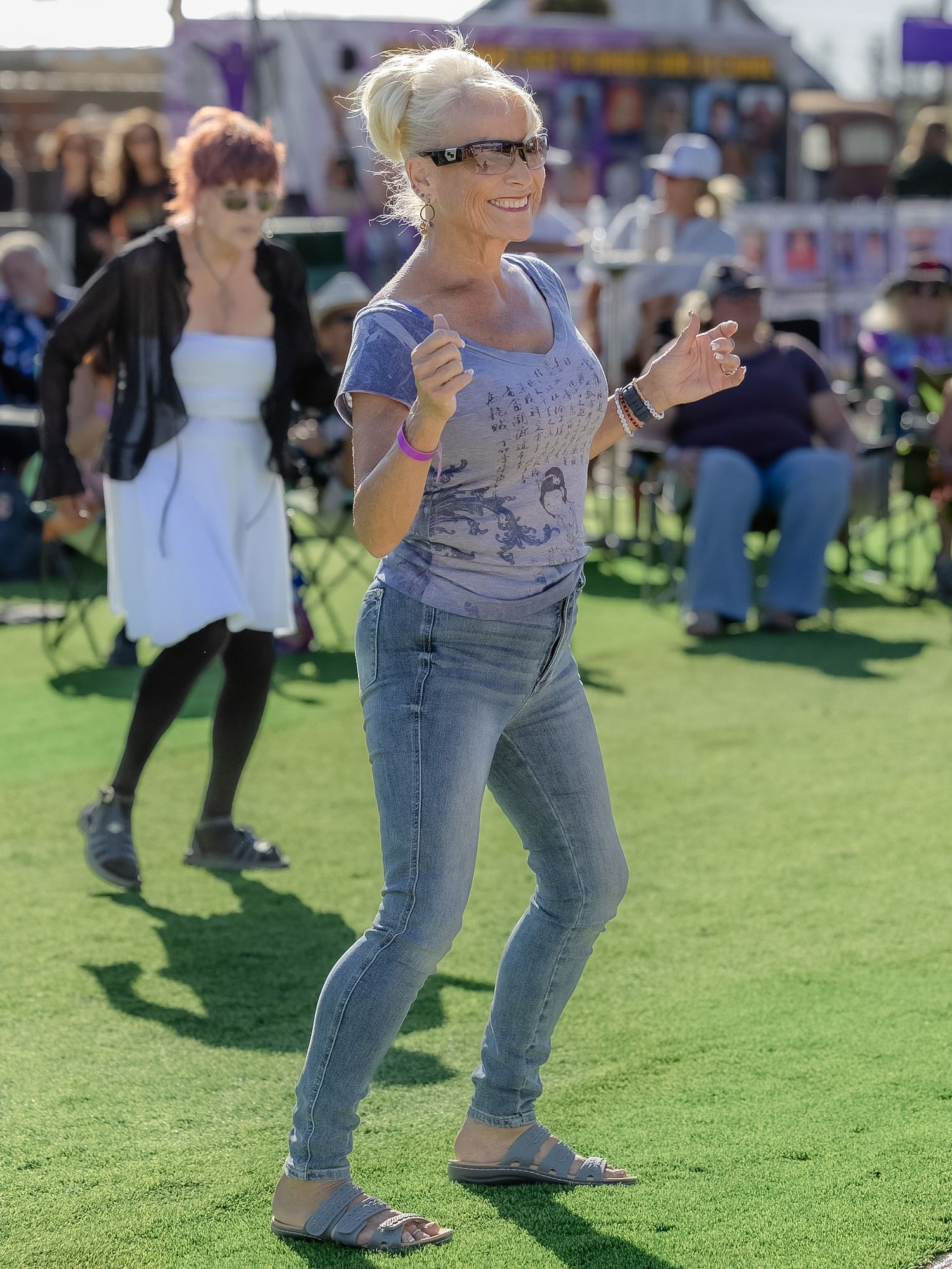 Blonde woman in jeans and gray t-shirt dancing on green grass at an outdoor music festival.