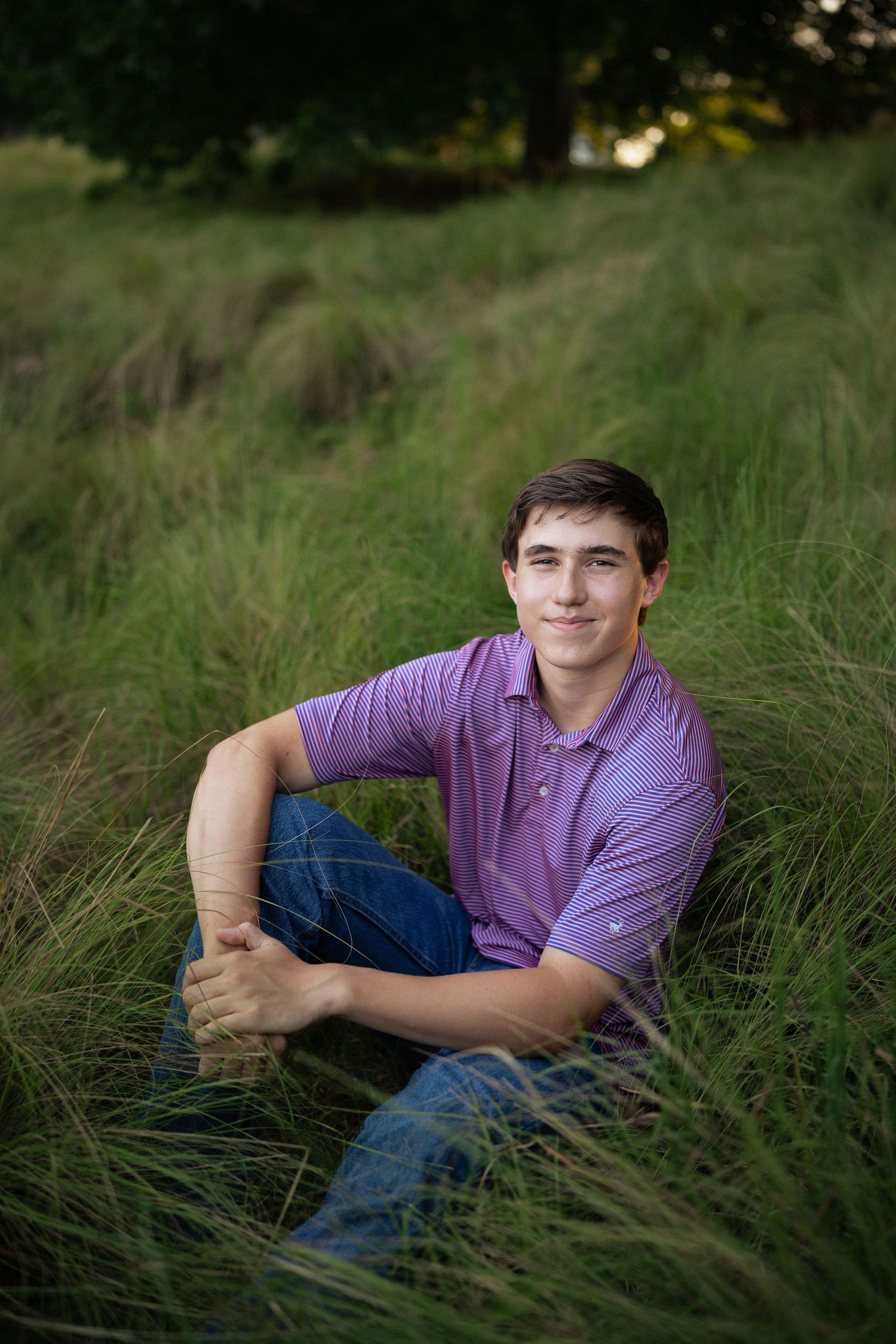 sitting in tall grass wearing a purple polo shirt at NCMA for senior photos