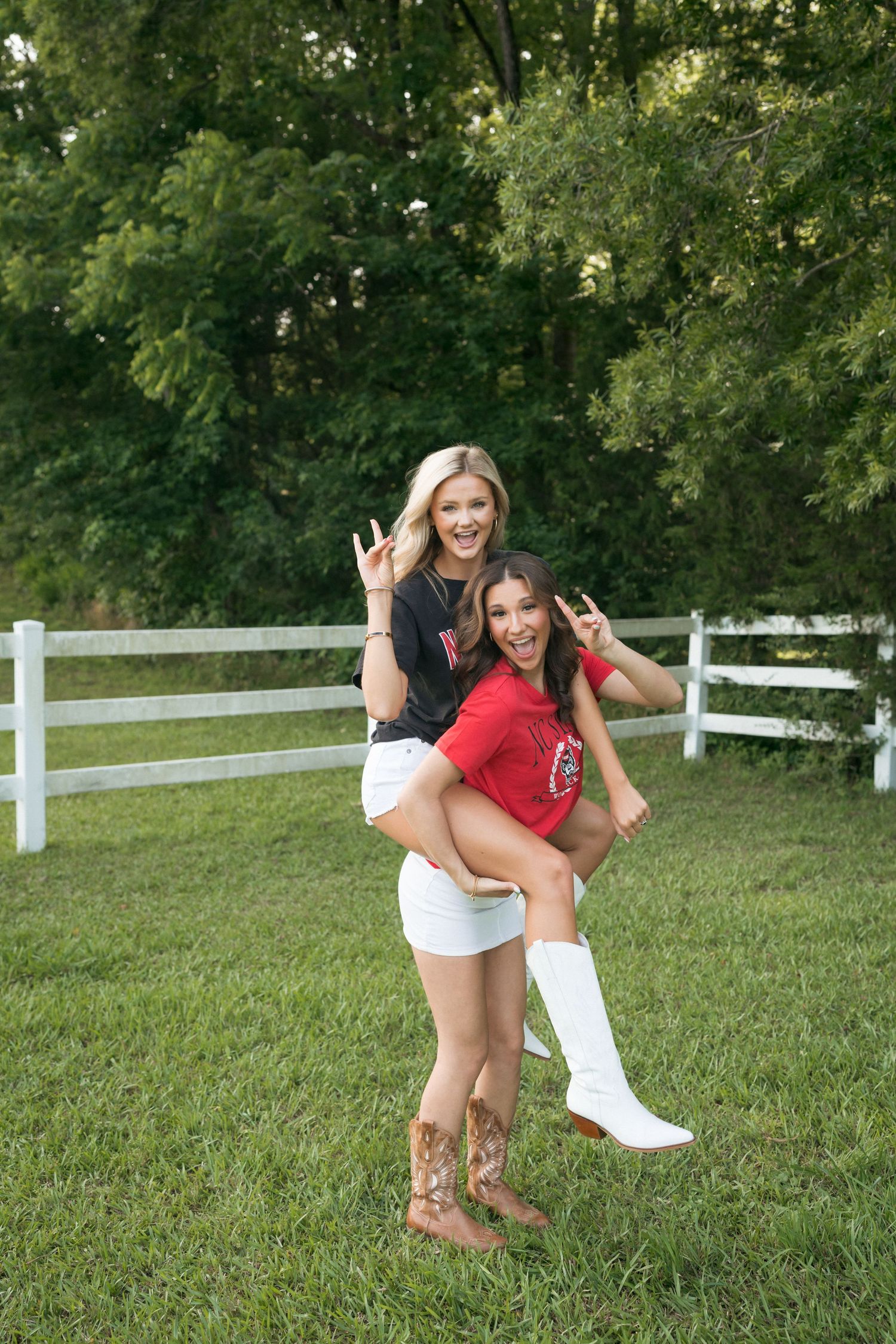 Two high school senior girls wearing NC State shirts doing a piggyback pose at Horseshoe Farm Preserve in Wake Forest, North Carolina.