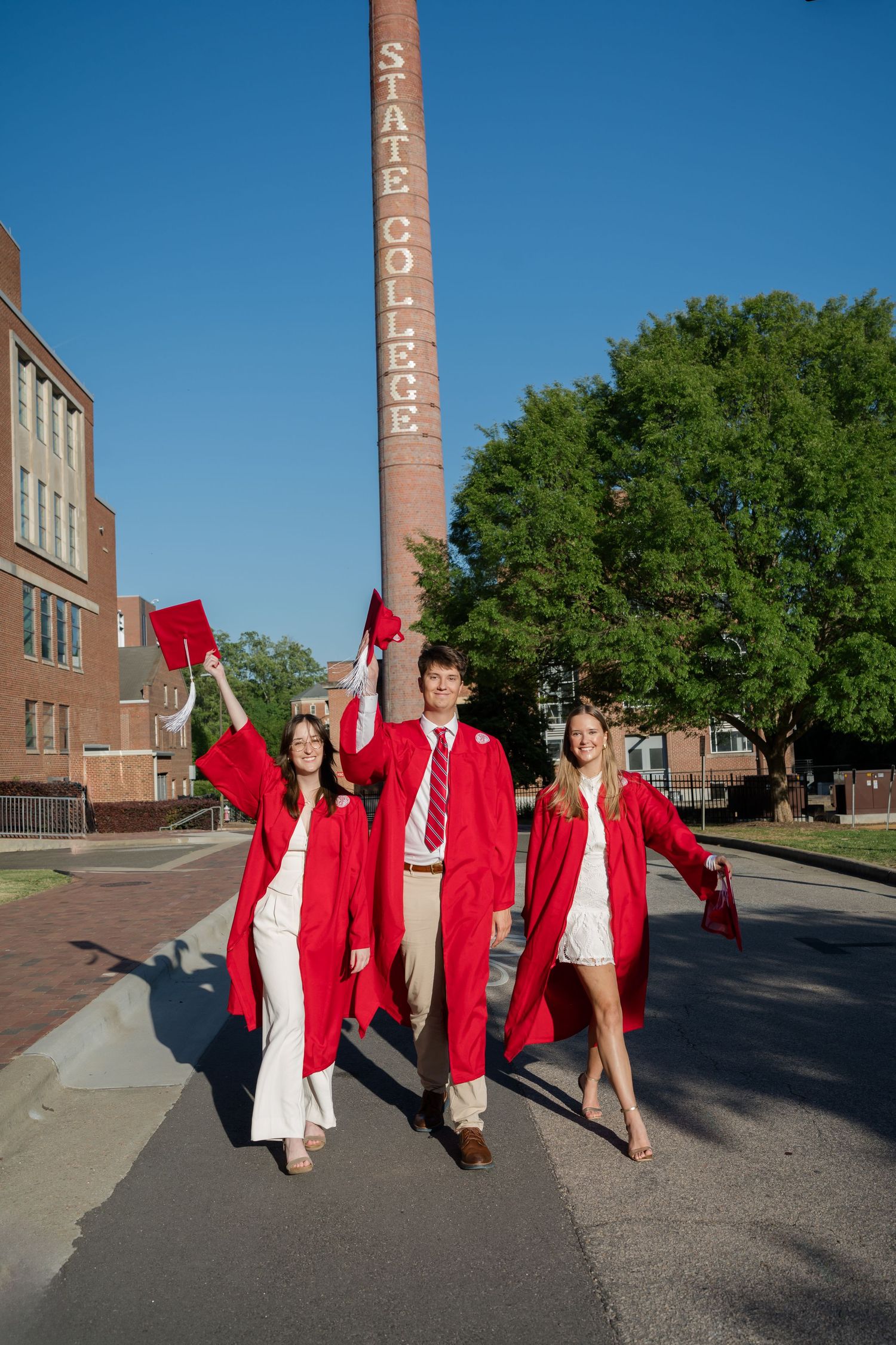 Triplet college graduates walking in cap and gown near the NC State smokestack on campus in Raleigh North Carolina