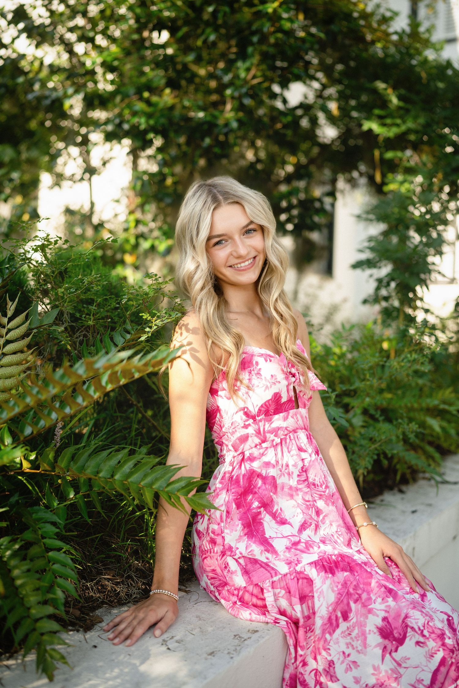 High school senior girl sitting on a stone wall with lush greenery in Wilmington, North Carolina during her senior portraits.