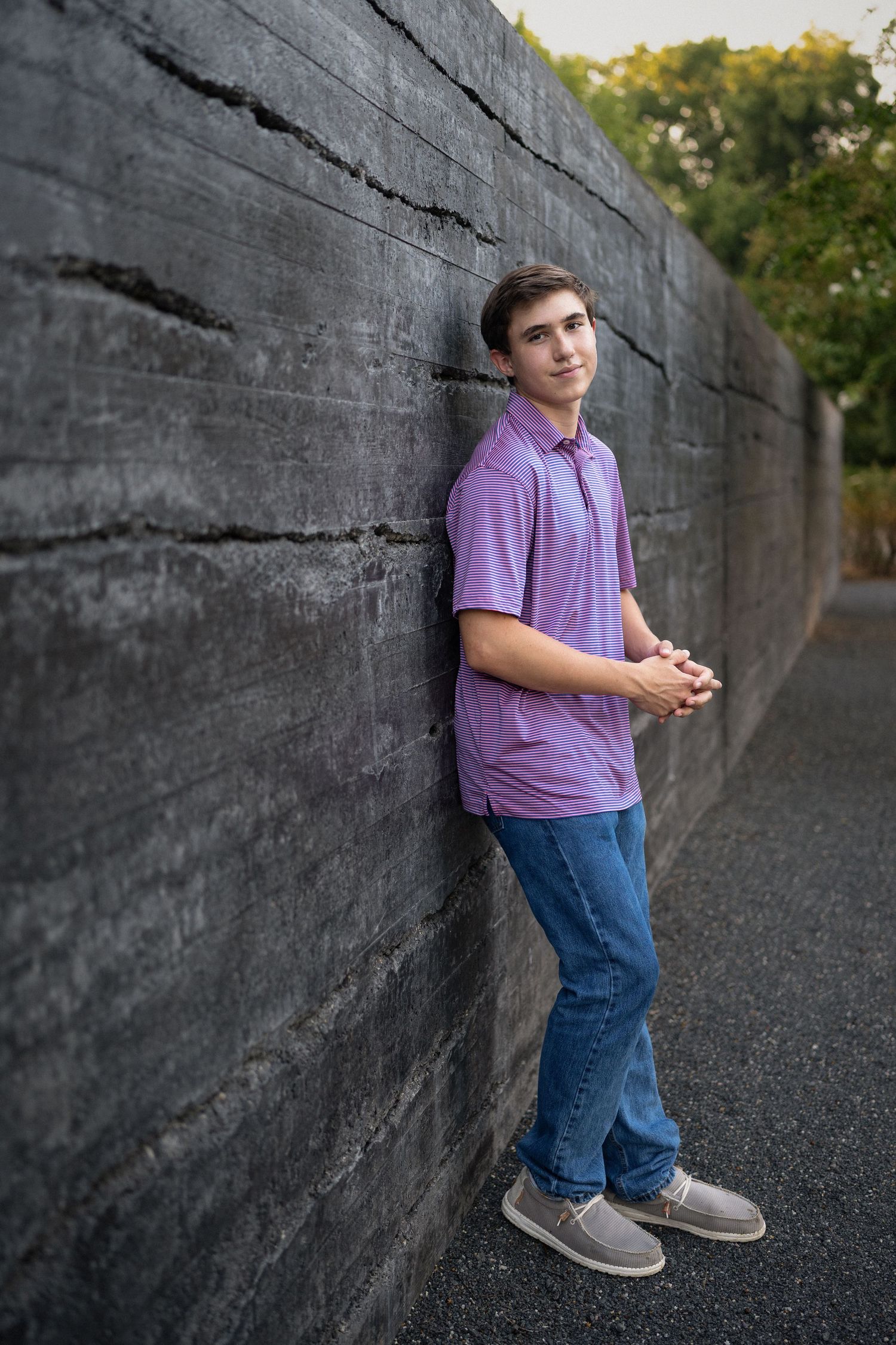 leaning against a stone wall in a purple polo shirt at NCMA for senior photos.