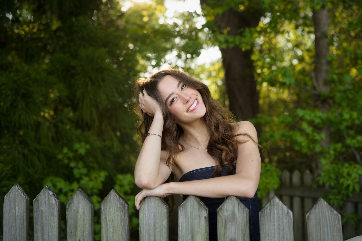 High school senior girl in a denim dress leaning on a fence at Horseshoe Farm Preserve in Wake Forest, North Carolina.