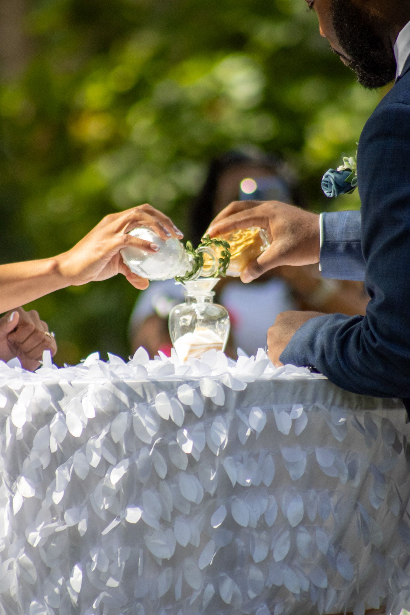 Hands exchange drinks across an elegant white textured table at an outdoor wedding reception.