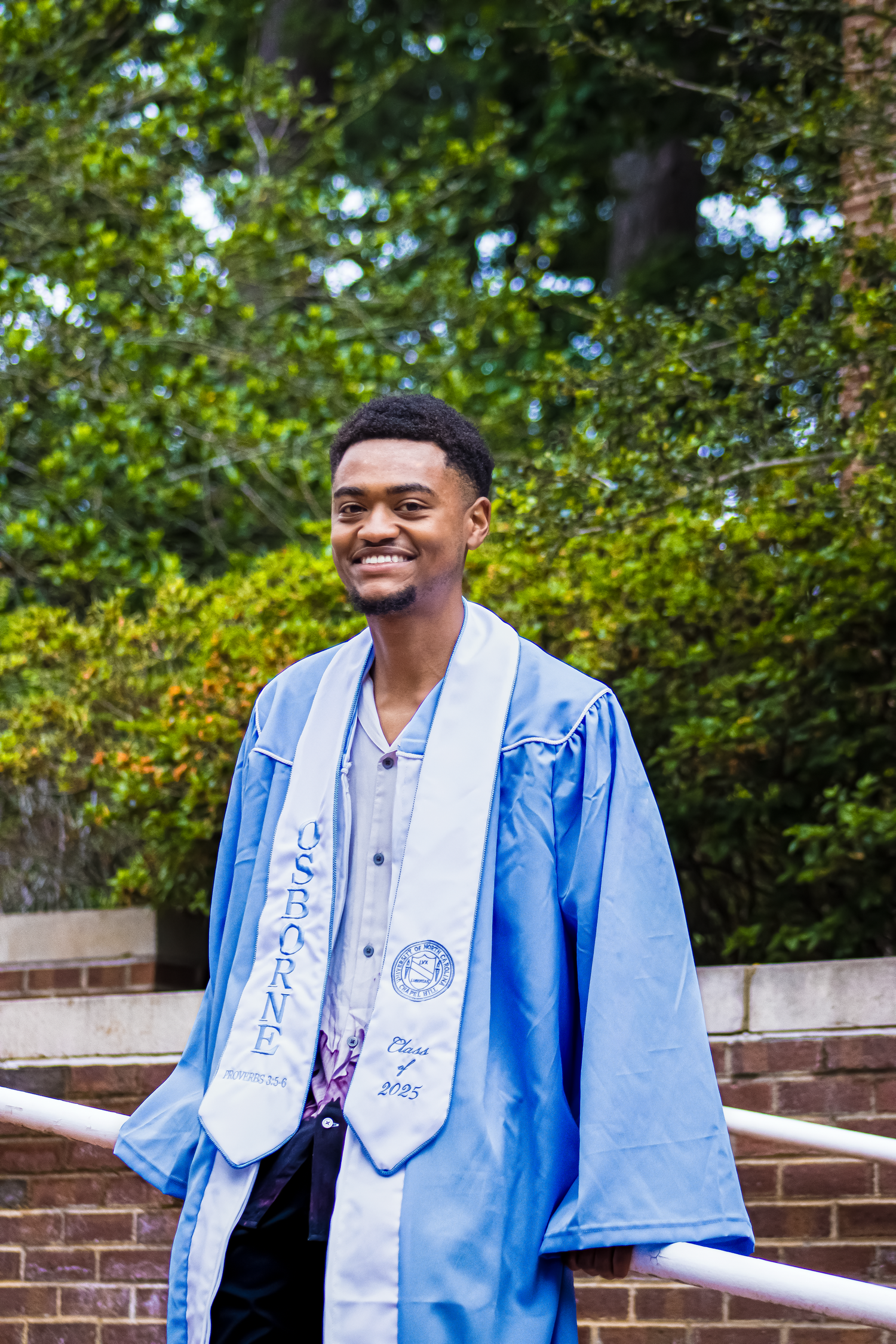 Smiling young man in light blue graduation gown and stole posing outdoors near brick wall and green trees.