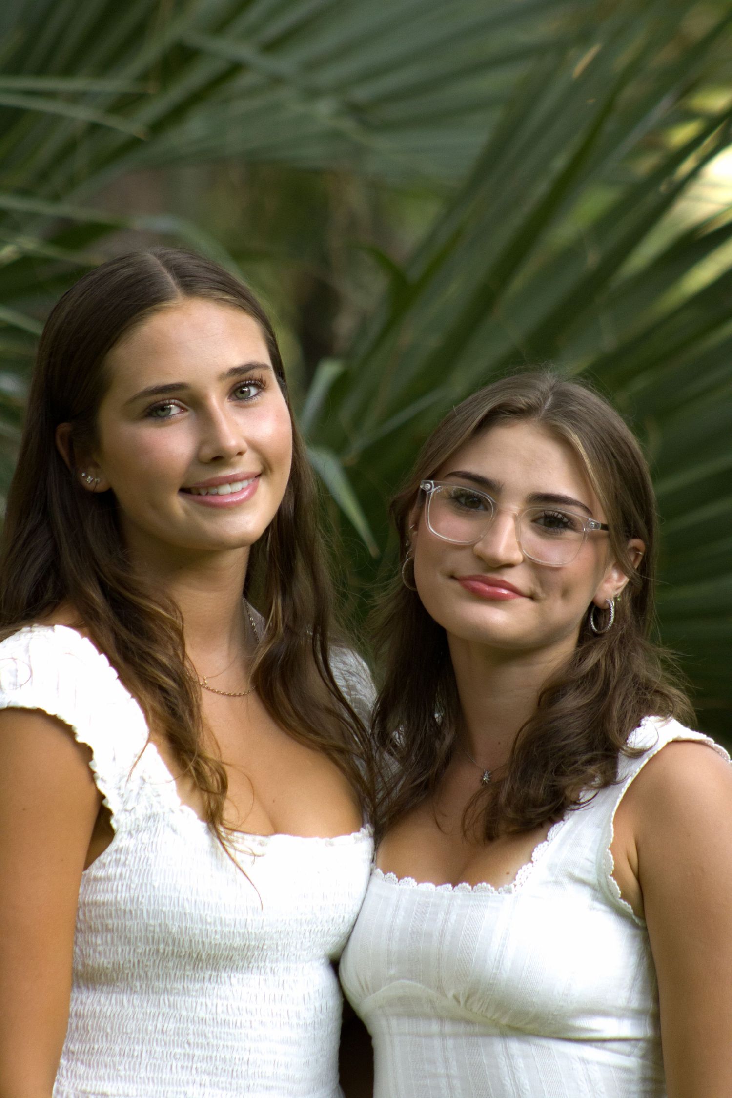 Two people in white tops pose together against a green palm frond background for a series of casual portraits.