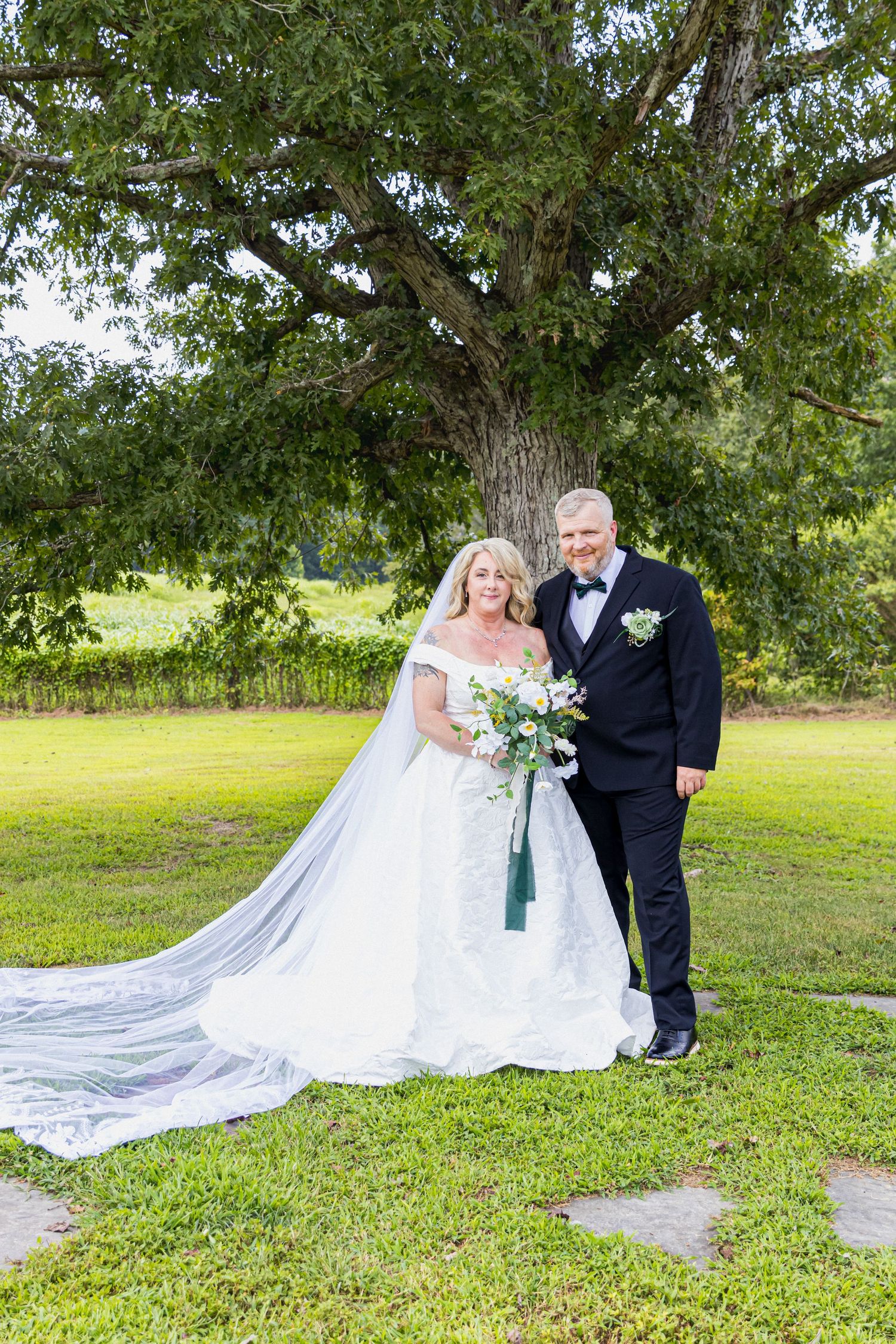 Bride and groom posing together under a large tree on green grass, bride in lace gown with veil, groom in black tuxedo.