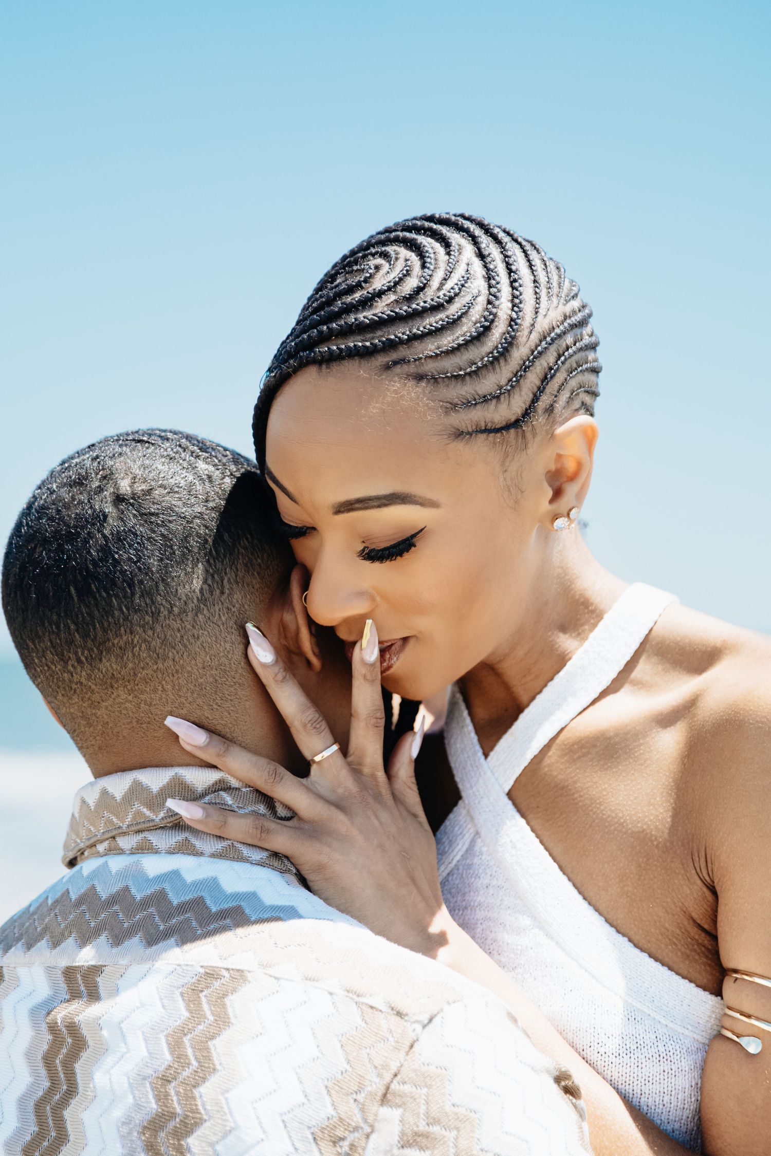 A couple sharing an intimate moment outdoors, woman with intricate cornrow braids gently touching mans face against blue sky.