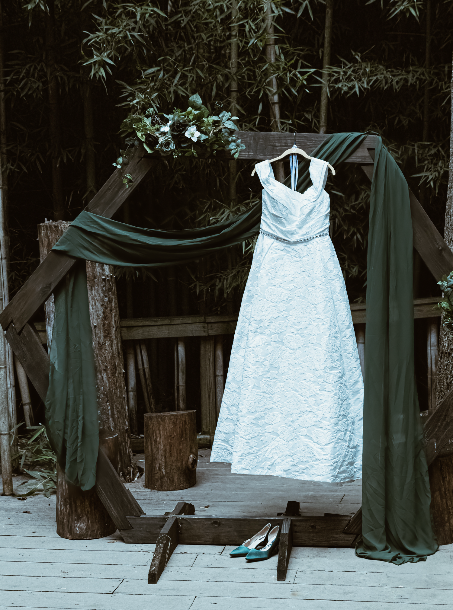 White wedding gown hanging on rustic wooden arch draped with dark green fabric, with teal heels below.