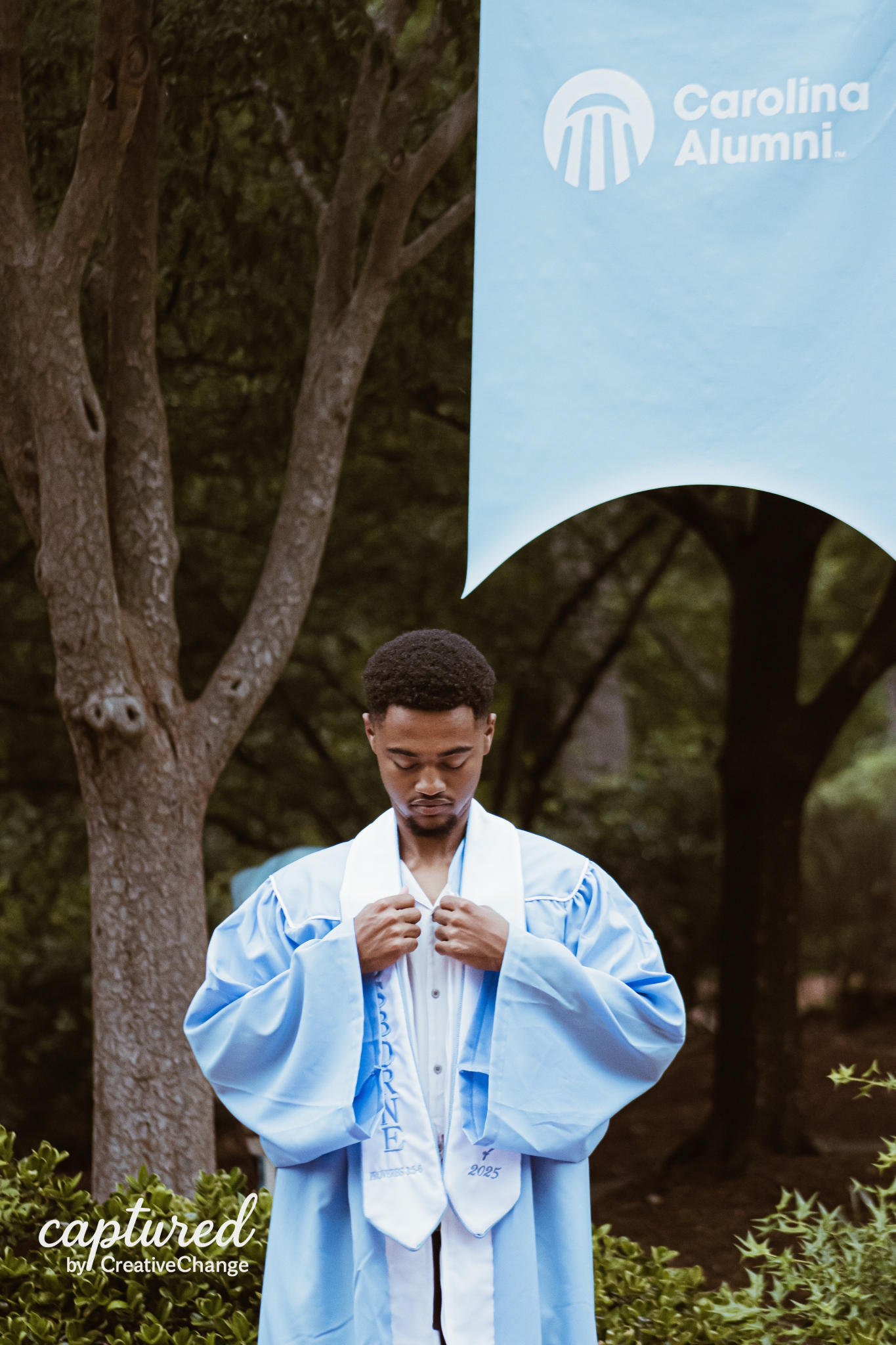 A young man in a light blue graduation gown adjusts his stole outdoors near a Carolina Alumni banner.