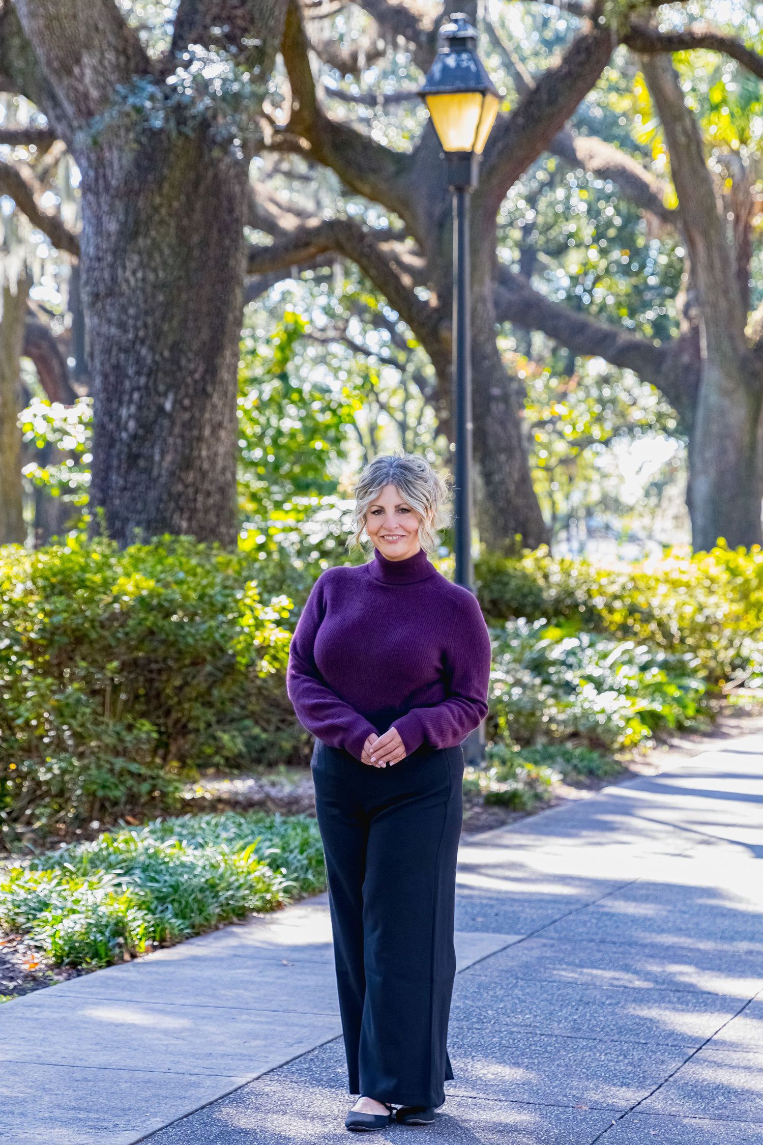 Smiling woman in purple turtleneck and black pants stands on a tree-lined park path beside a vintage lamp post.