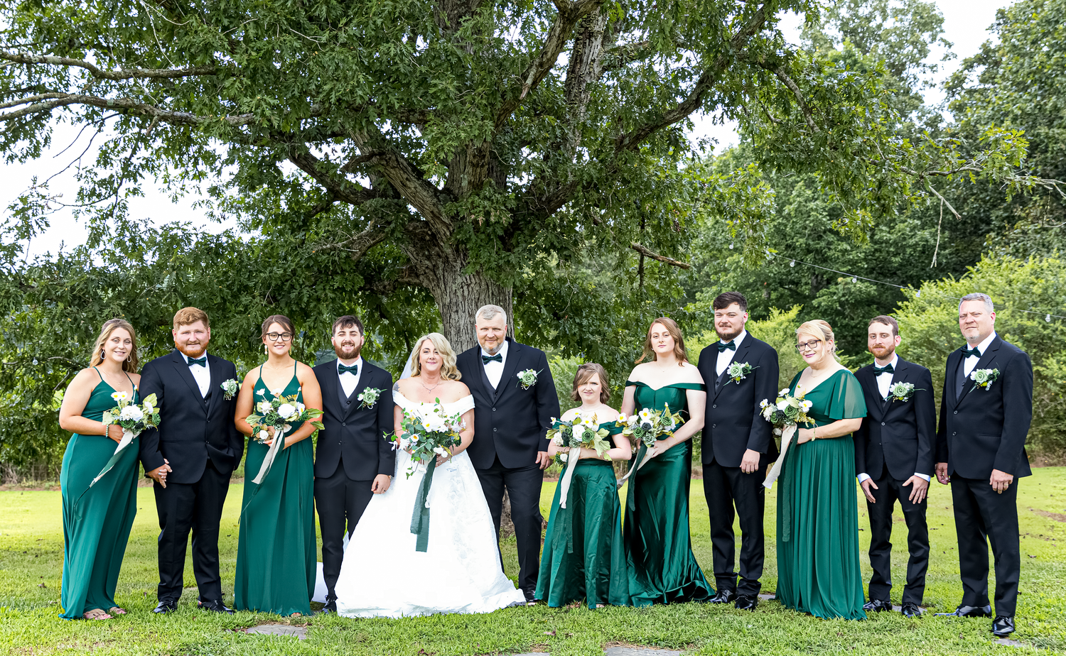 Wedding party in emerald green dresses and navy suits poses together on grassy lawn during outdoor celebration.