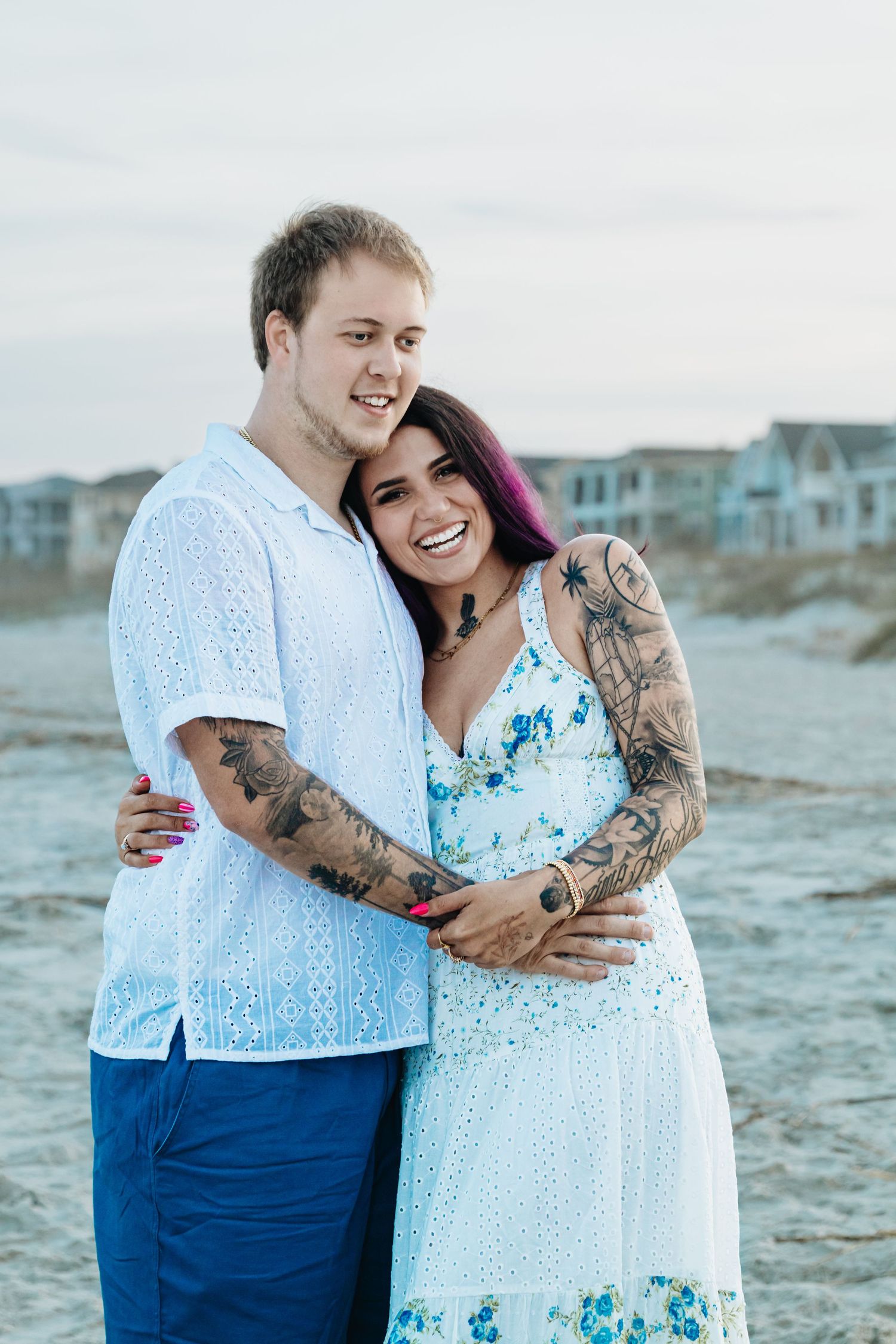 A smiling couple with tattoos embraces on a beach, wearing white and blue outfits with colorful houses in the background.