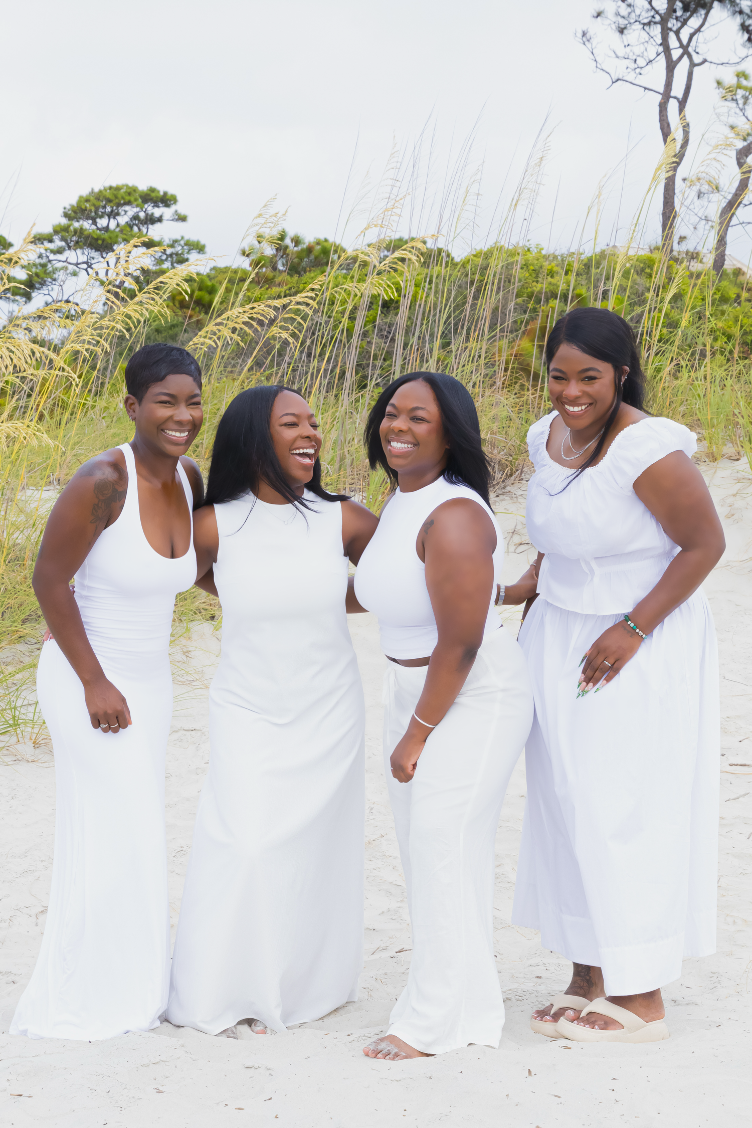 A group of people wearing white dresses pose together joyfully on a beach with trees in the background.