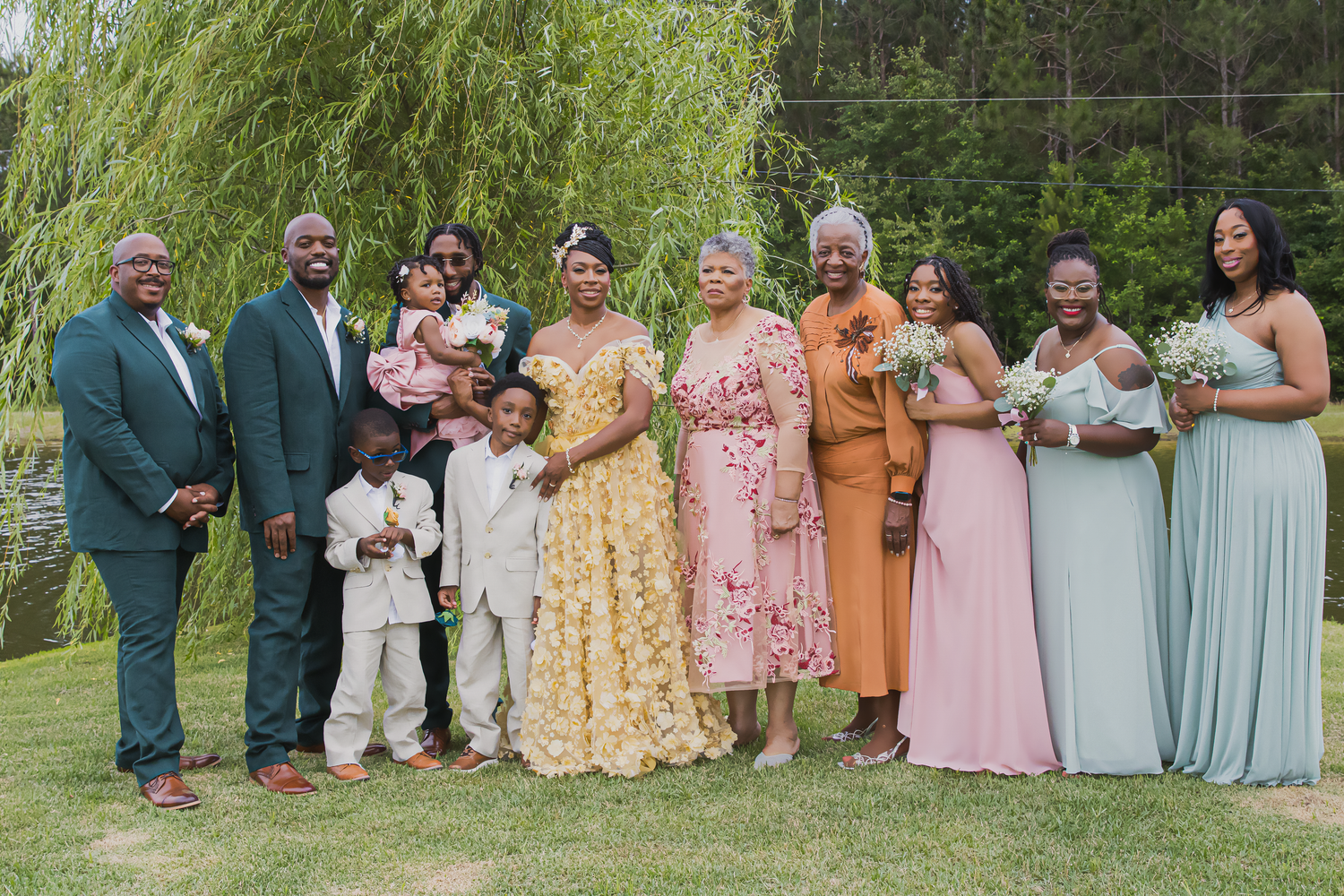 Wedding party members pose together in pastel colored dresses and suits on a lush green lawn.