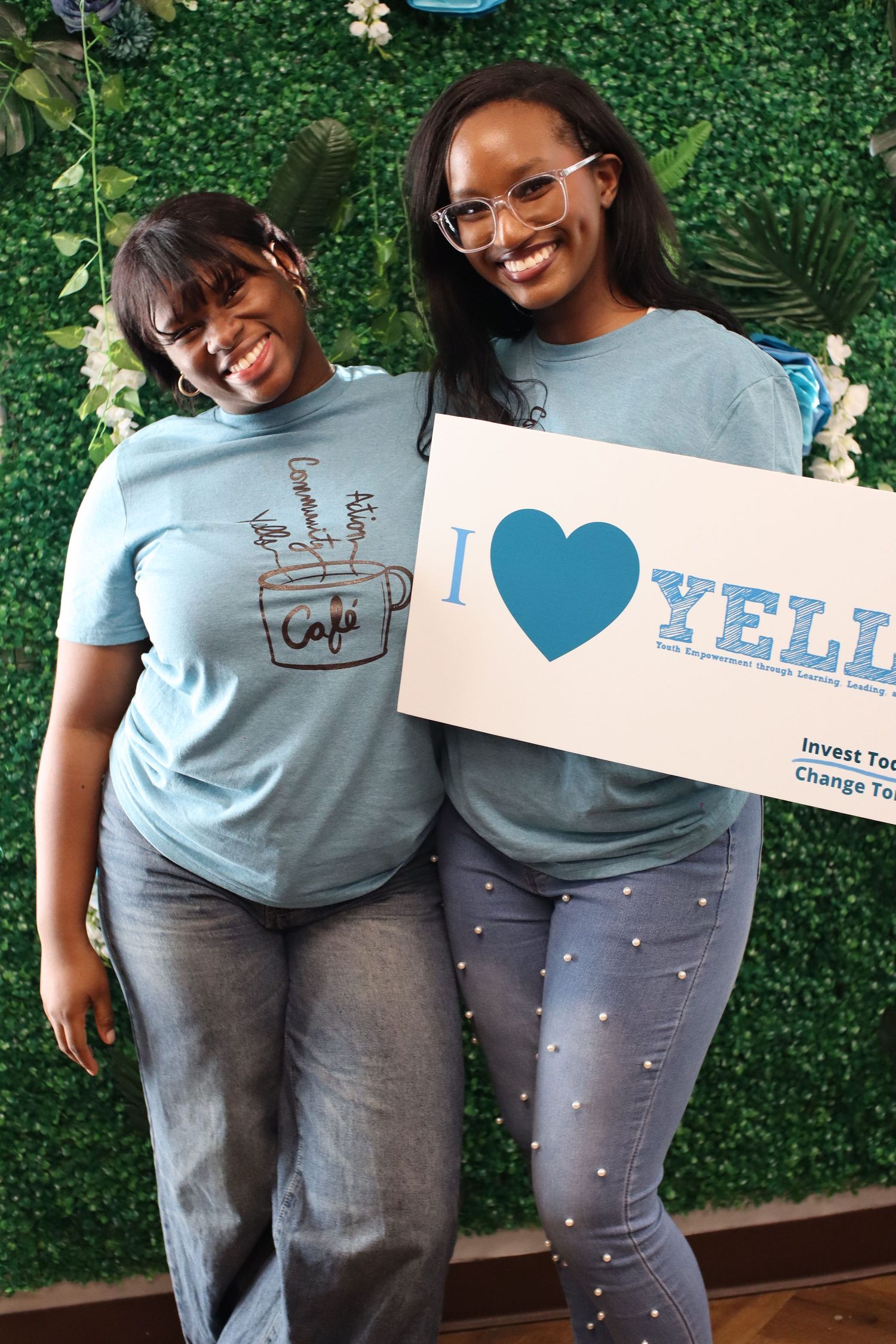 Two women in matching light blue cafe t-shirts smile while holding an I Love YELL sign in front of a green floral wall.
